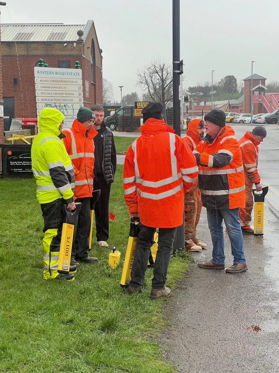 Group of people in high visibility safety jackets and gear gathered on a wet sidewalk next to a street, with a sign for Western Road Estate and a parking lot in the background.