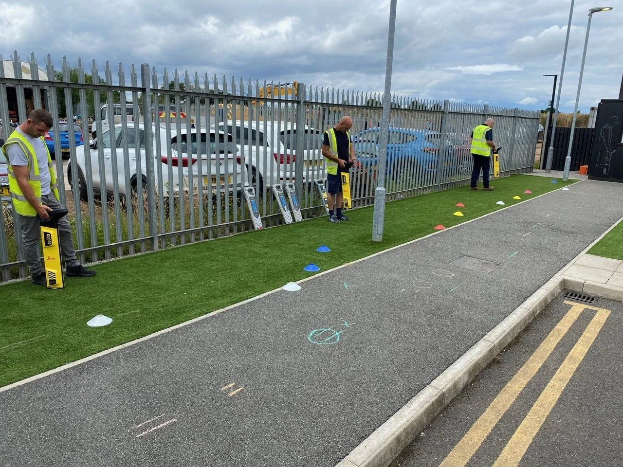 Three men in yellow safety vests practicing driving or parking electric scooters on a small artificial grass track with cones in an outdoor parking lot.
