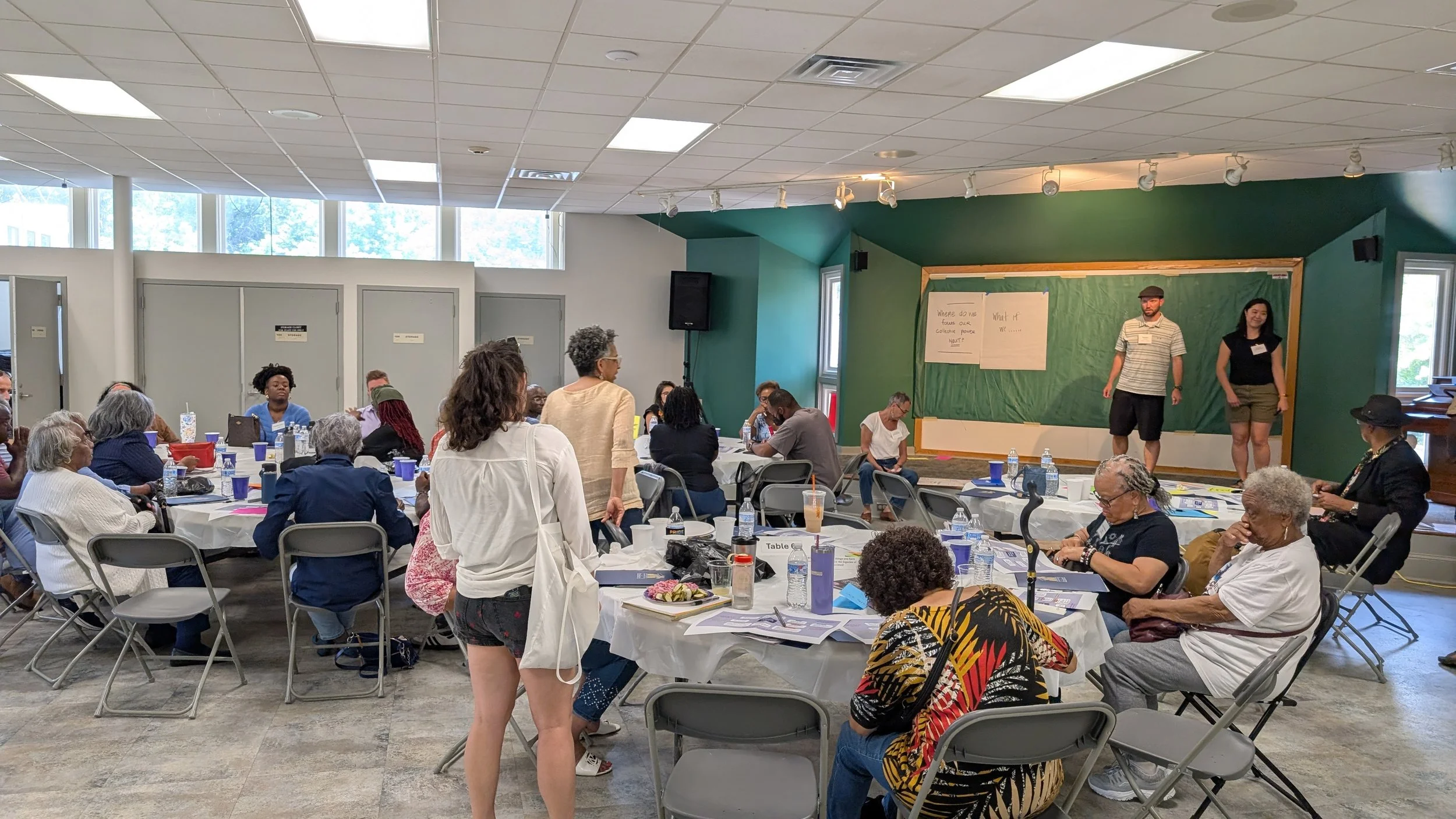 A diverse group of people gathered in a conference room, seated around round tables with white tablecloths, drinking water and coffee, facing a stage with two presenters standing in front of a green chalkboard with large sheets of paper attached. One woman is standing, and another man is walking toward the stage. The room is well-lit with natural light from windows and ceiling lights.