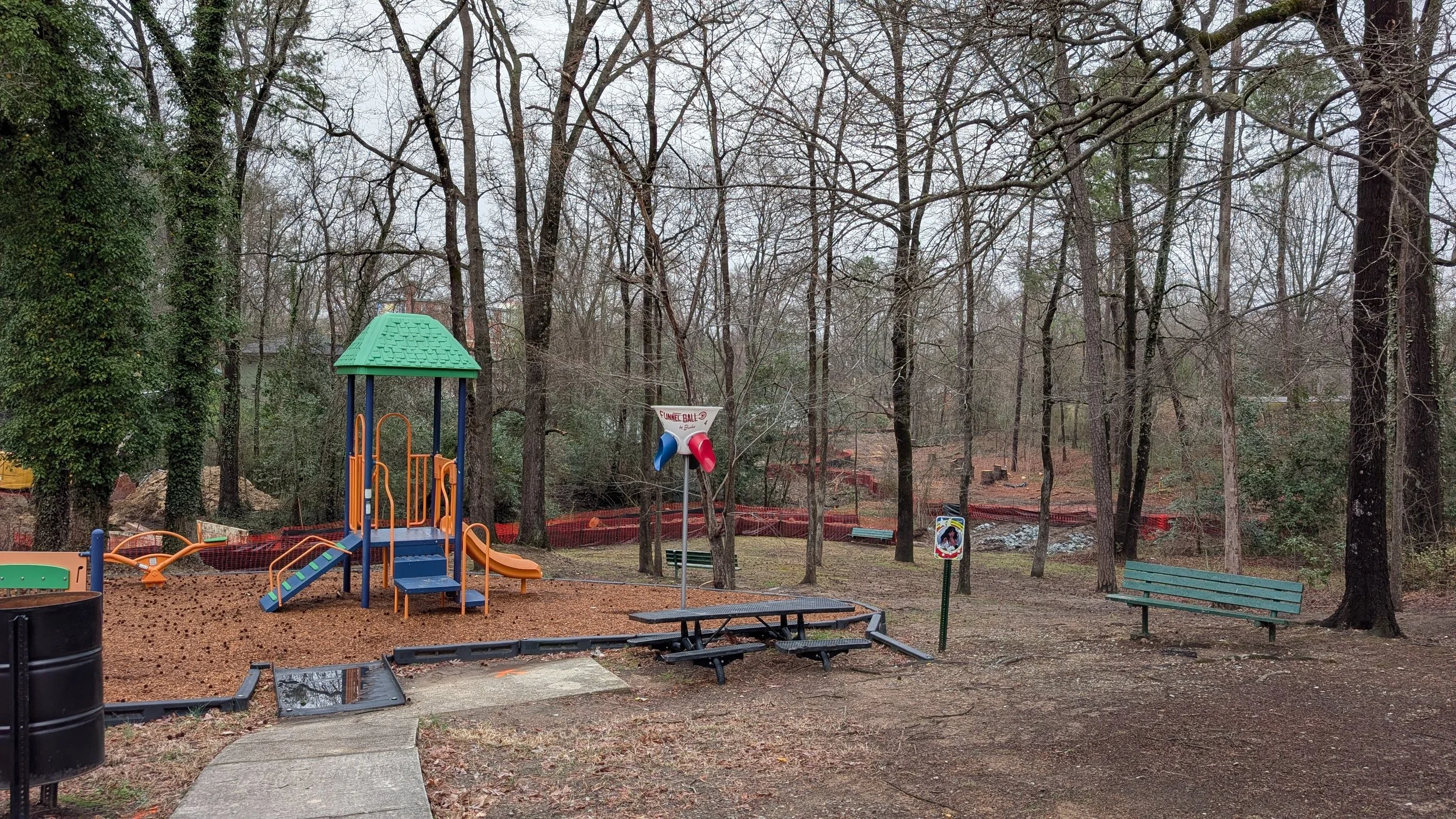 Empty playground with a blue and orange play structure, a green bench, a sign for a funnel ball game, and tall leafless trees
