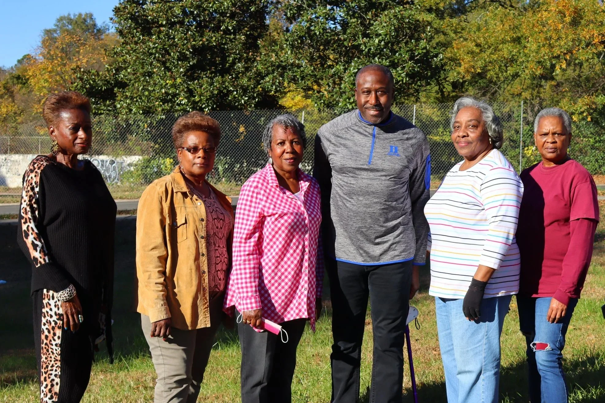 Group of seven diverse older adults standing outdoors on grass, with trees and a fence in the background, smiling for a photo.