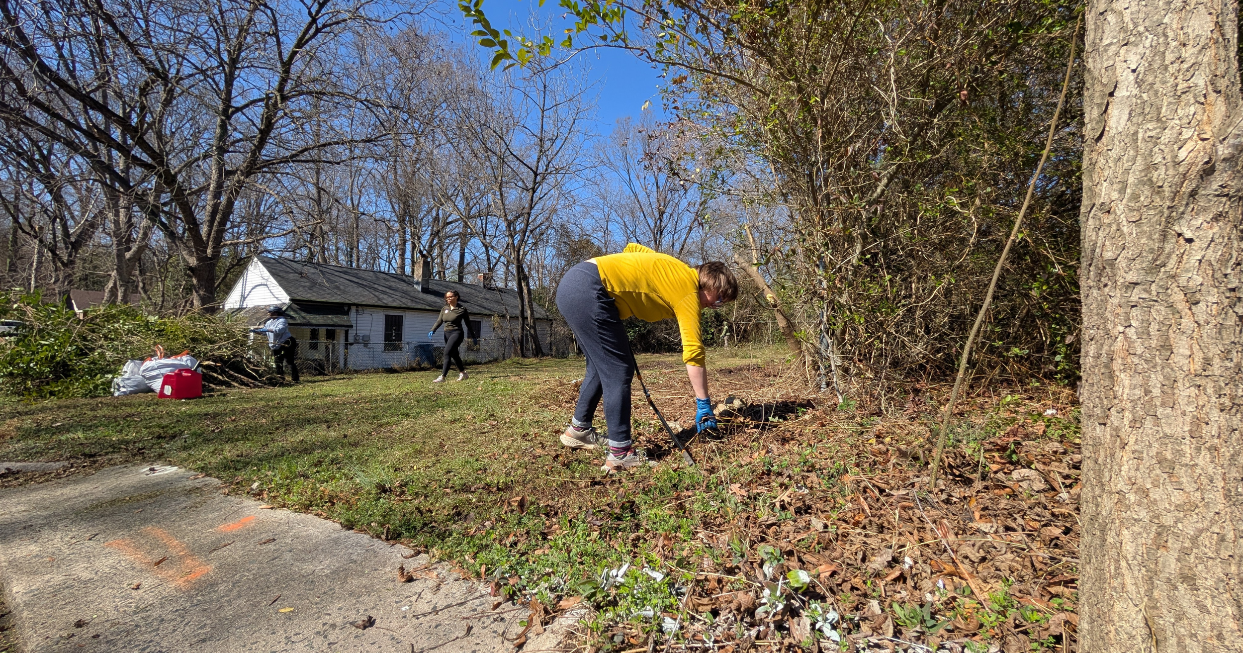 People working outdoors in a yard, with one person using a rake to clear leaves and debris, and others further away near a pile of branches, during a sunny day with leafless trees and a house in the background.