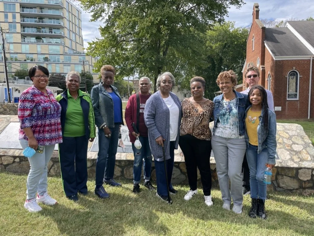 A group of ten diverse women and two men standing outdoors on a grassy area in front of a stone monument, with trees, a brick building, and a modern high-rise building in the background on a clear day.