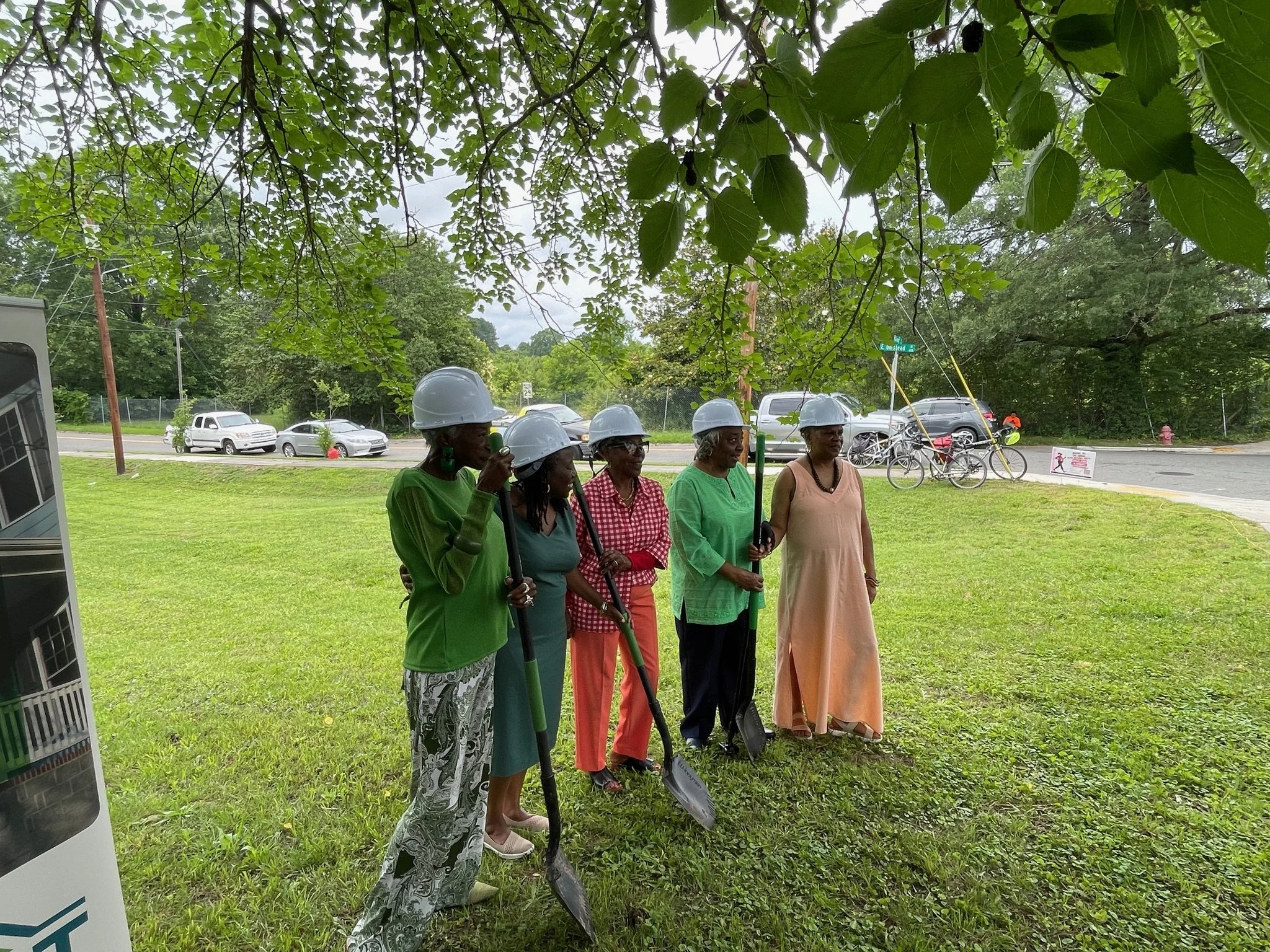 Five women standing outdoors under tree branches, wearing construction helmets and holding shovels, in a grassy area near a street with parked cars and bicycles.