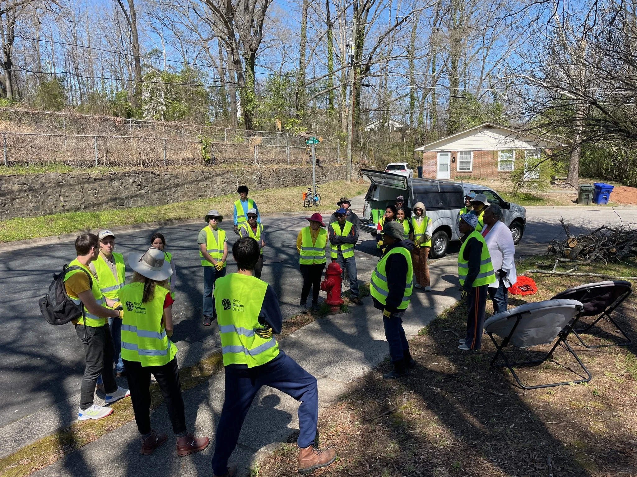Group of people wearing yellow safety vests gathered outdoors on a street, listening to a speaker during a community cleanup or event on a sunny day.