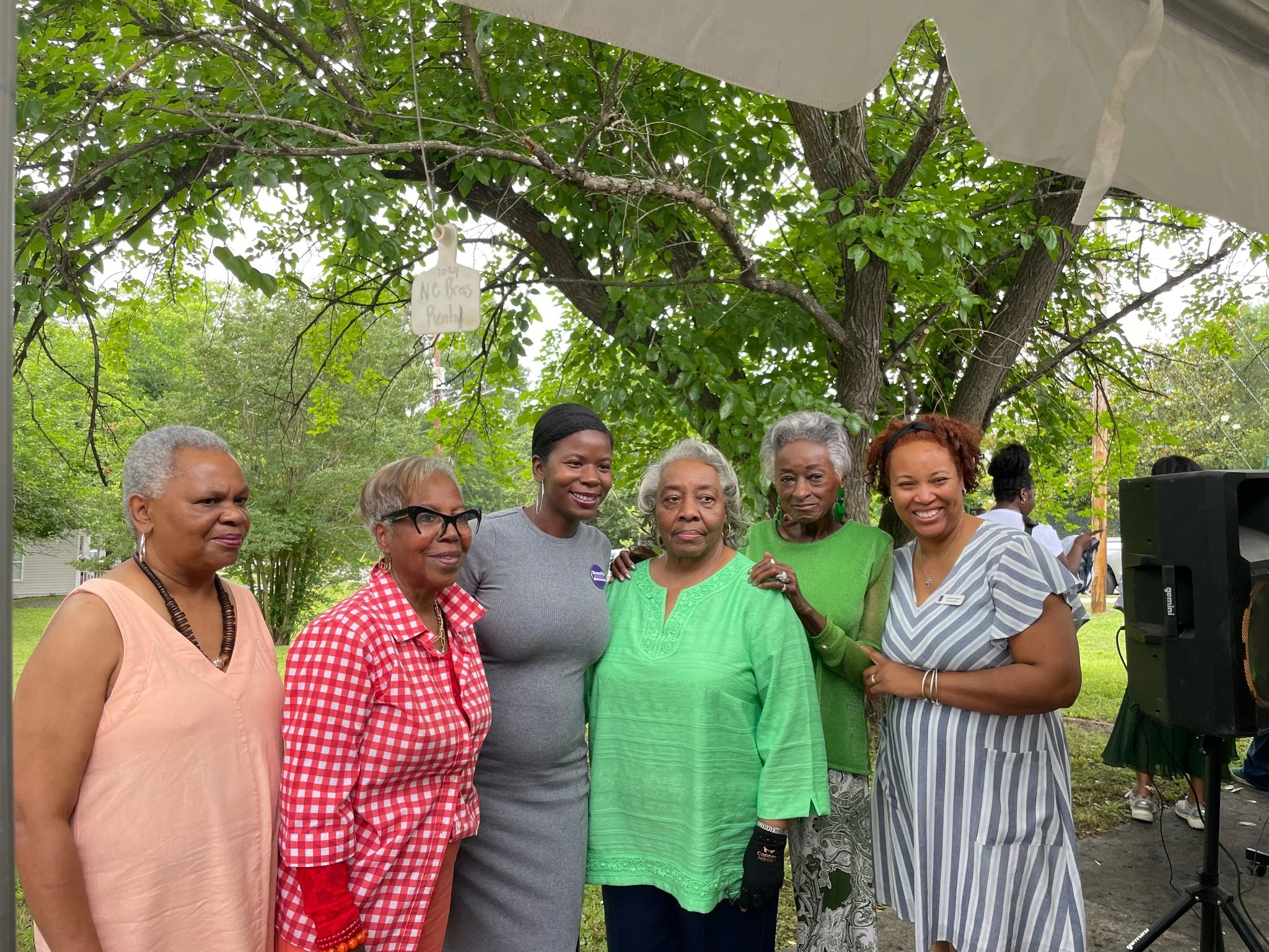 Six women standing outdoors in front of a large tree, smiling for a photo. There is a white tent and speaker nearby, suggesting a gathering or event.