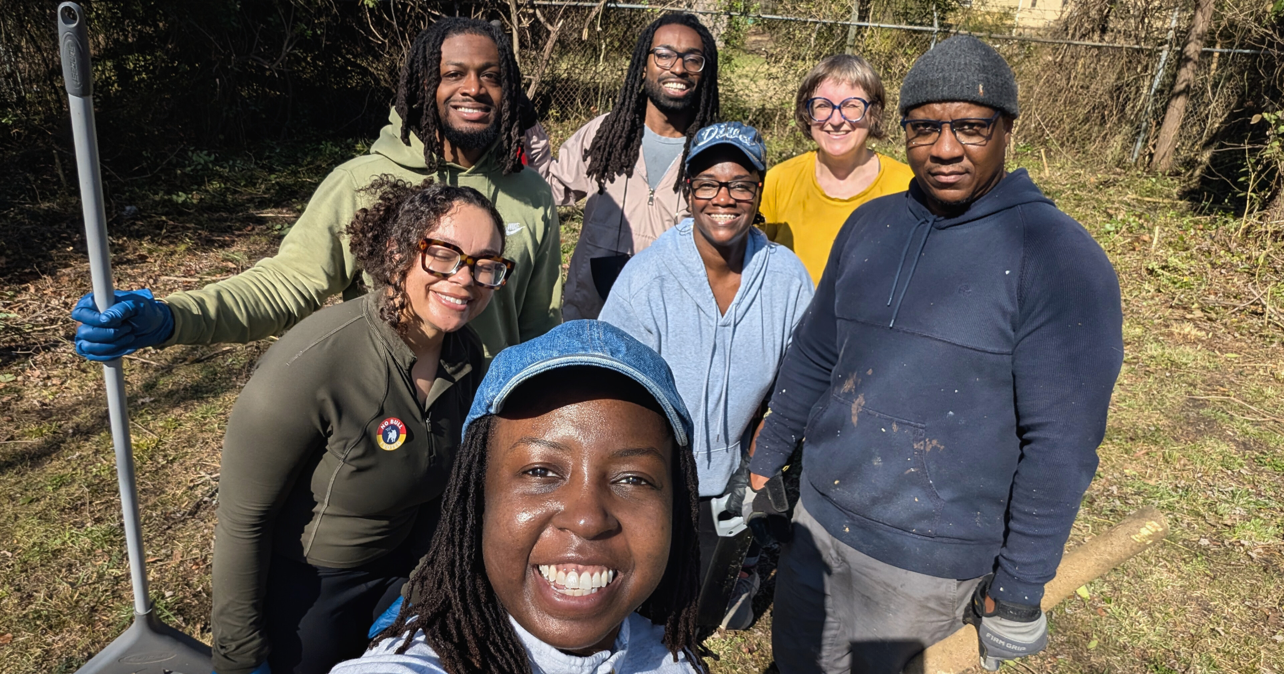 Group of eight people smiling outdoors, some holding tools for planting or gardening, in a natural setting with trees and grass.