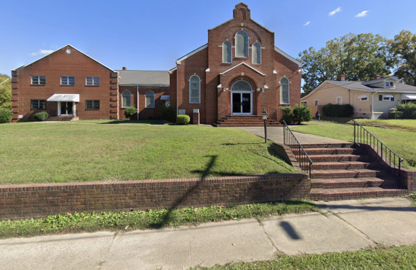 A brick church with steps leading up to the entrance, surrounded by a grassy lawn and neighboring houses, under a clear blue sky.