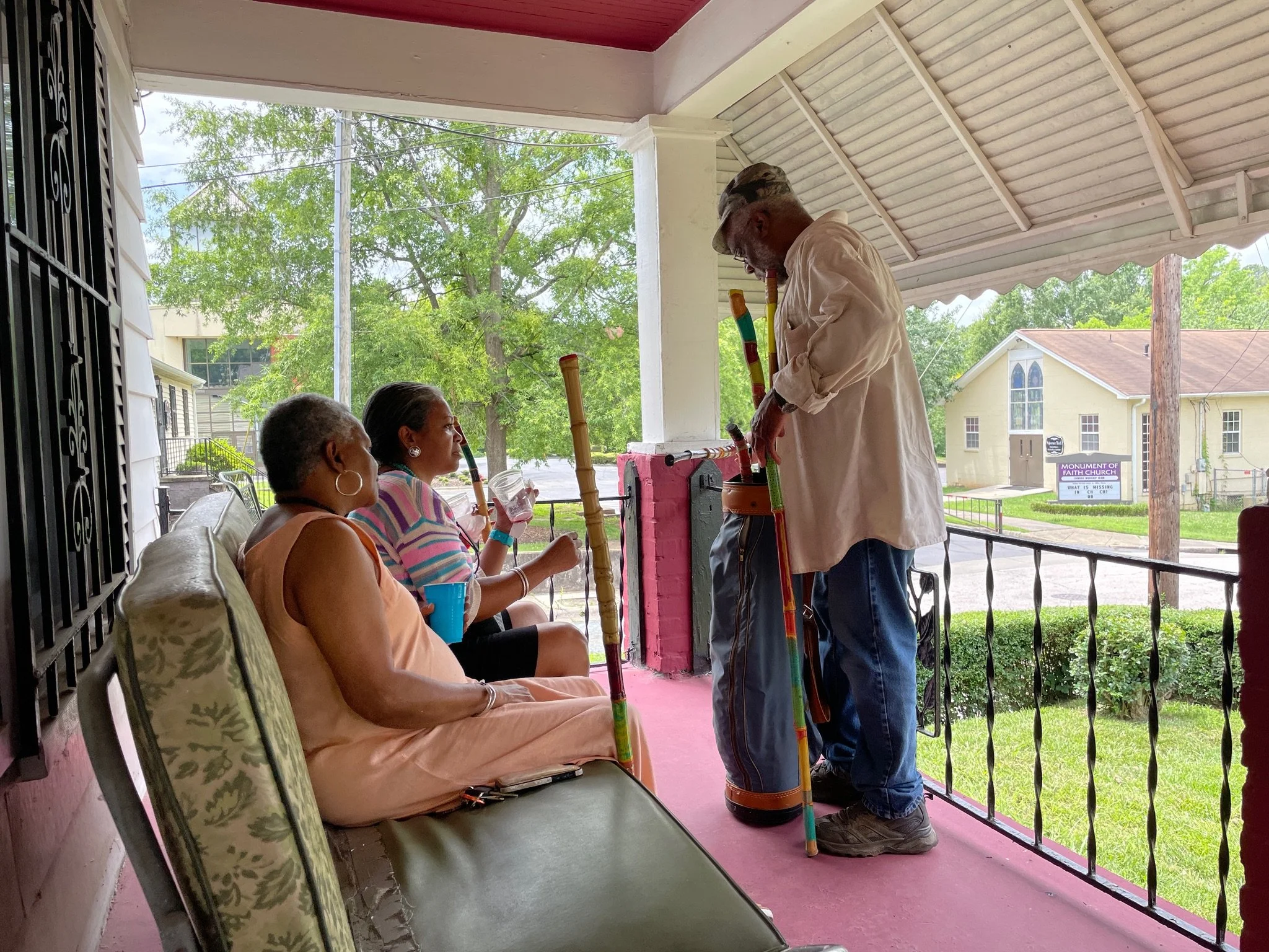 Three women sitting on a porch, one woman is playing a didgeridoo, and a man is standing, playing a didgeridoo, with tall, colorful wooden sticks around him.