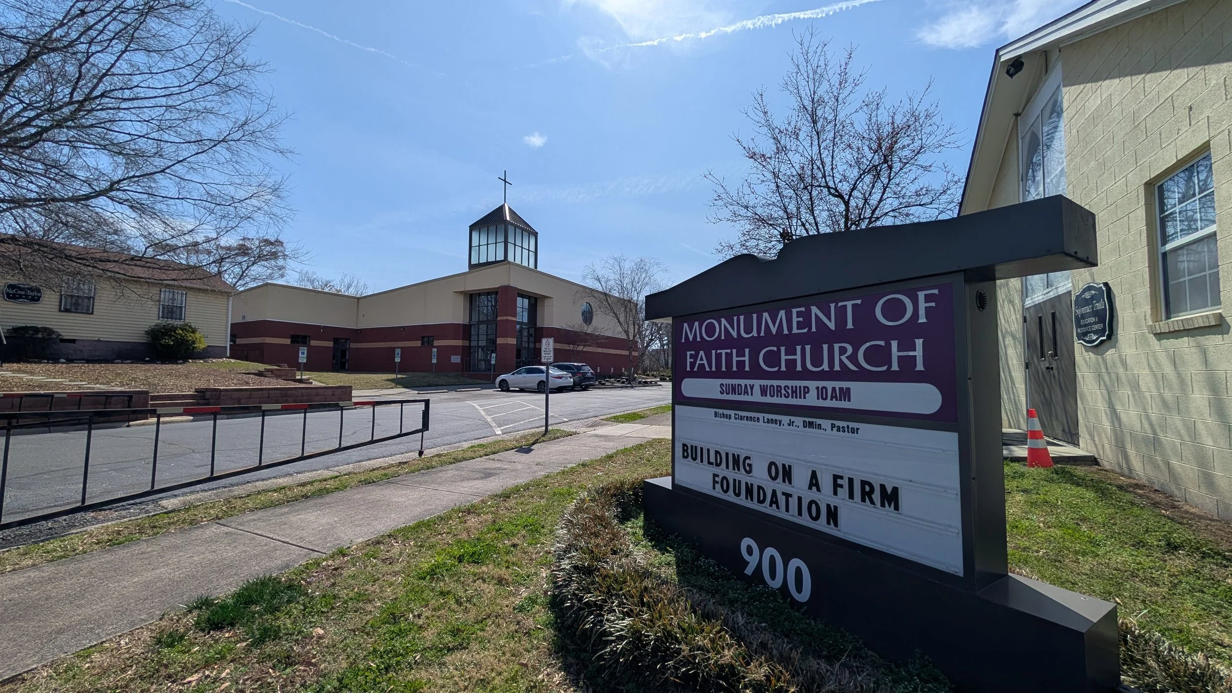Sign outside Parliament of Faith Church displaying service times and message, with church building, parking lot, and blue sky in the background.