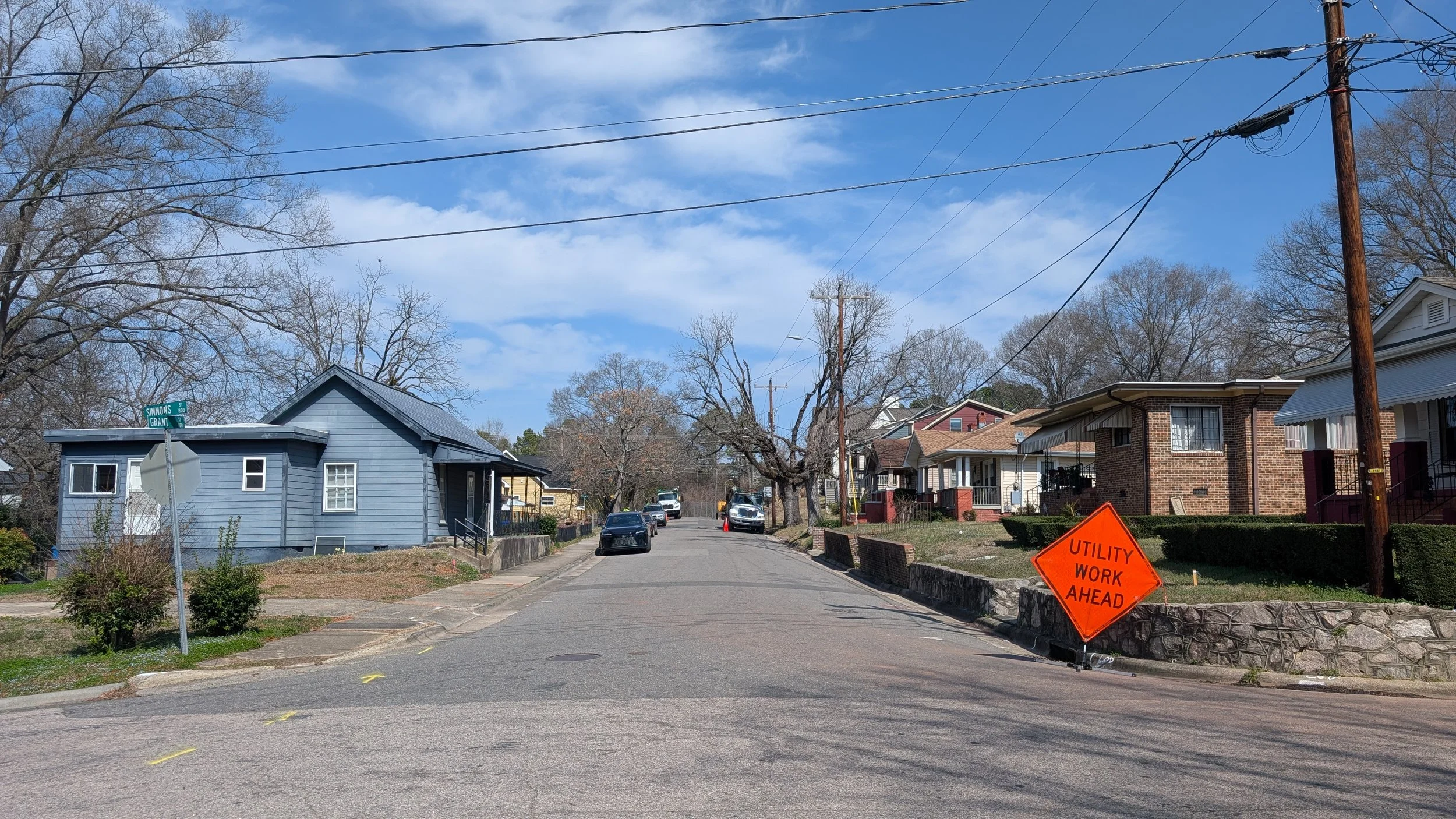 Residential street with houses, parked cars, utility lines, and a bright orange sign reading 'Utility Work Ahead'