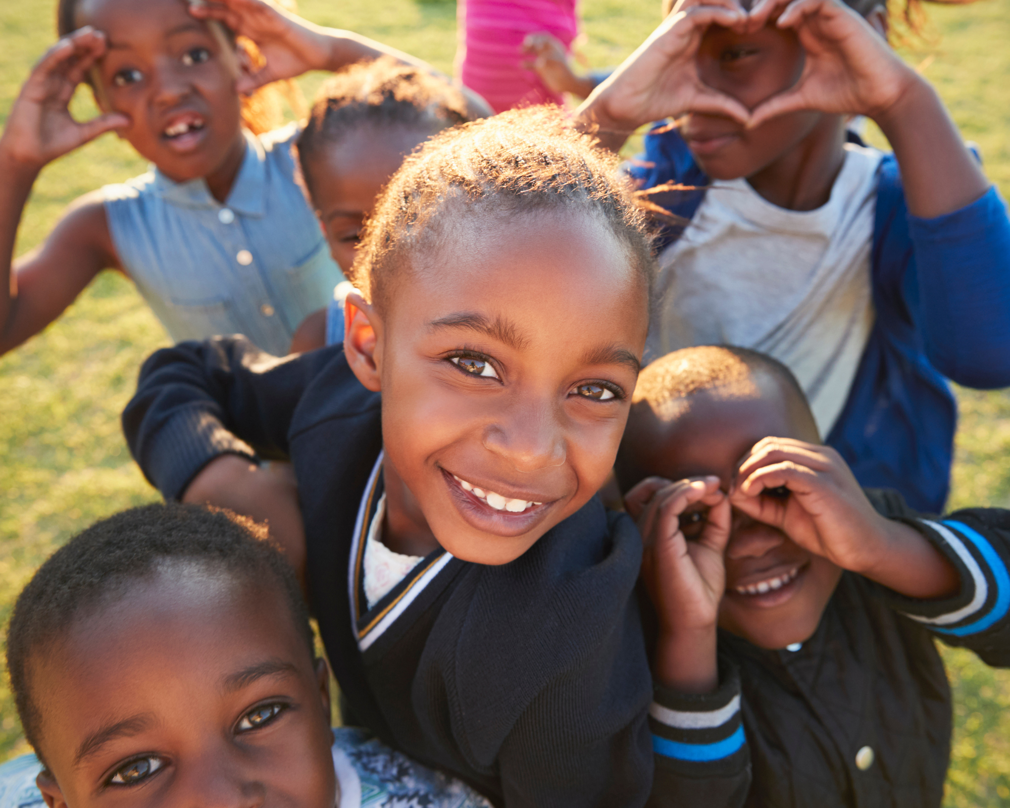 Smiling children playing outside, making heart shapes with their hands, on a grassy field during daytime.