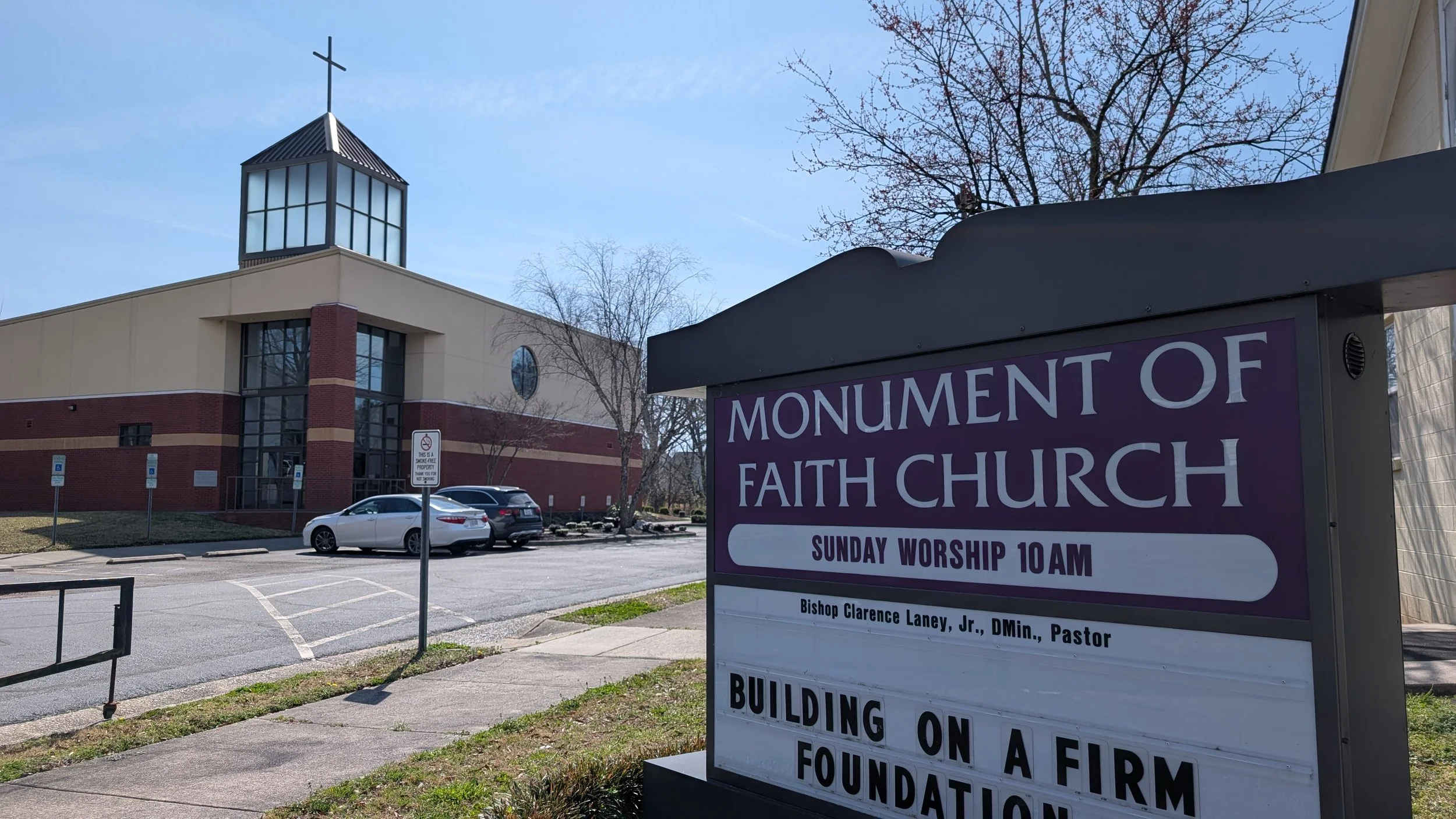 Exterior view of Monument of Faith Church building with a sign advertising Sunday worship at 10 am, led by Pastor Bishop Clarence Laney Jr. The building has a modern design with large windows and a cross on top, and a parking lot with several parked cars.