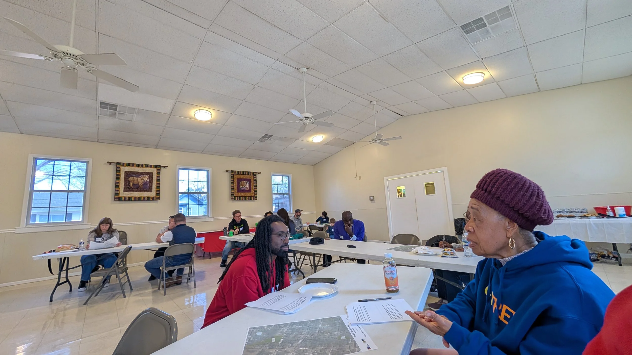 People sitting at tables in a large, well-lit room with beige walls and windows, some reading papers, engaging in conversation, or listening, with food and drinks on the tables.