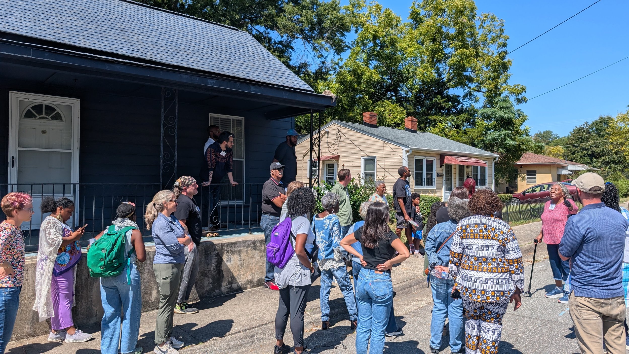 A diverse group of people gathered outside a house, listening as a woman speaks. The house has a porch, dark blue siding, and a gray roof. Some attendees have backpacks and are casually dressed on a sunny day.