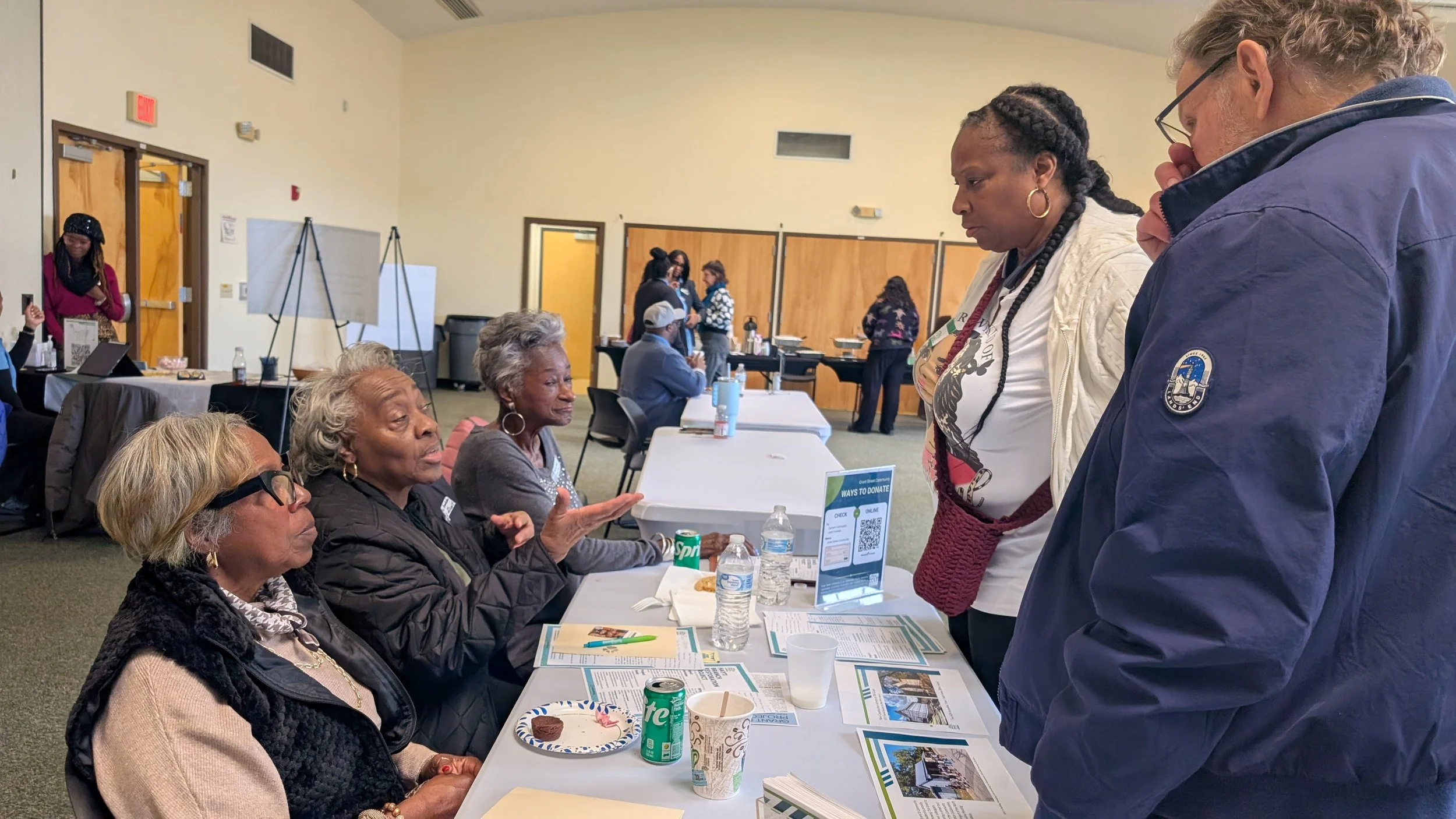 A group of elderly women and a man engaging in conversation at a community event with informational materials and drinks on the table.