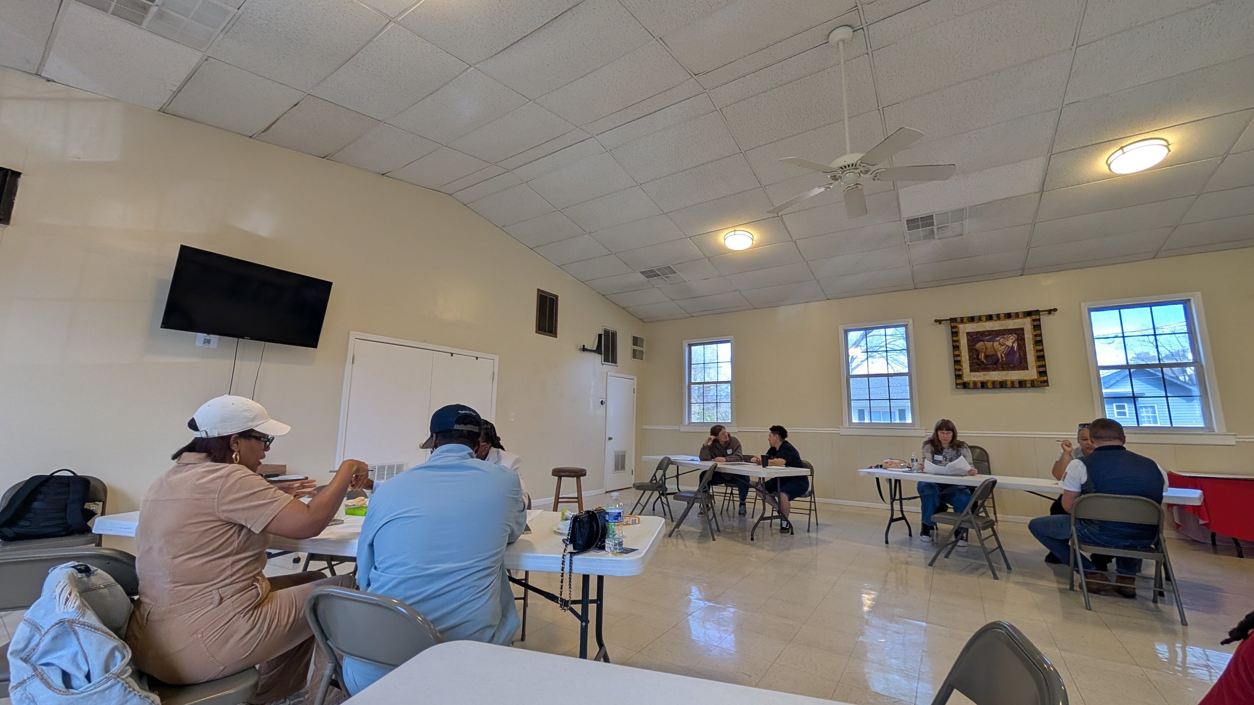 People sitting at tables in a room with beige walls, windows, a wall hanging, and a television. Some are talking, some are eating or drinking.