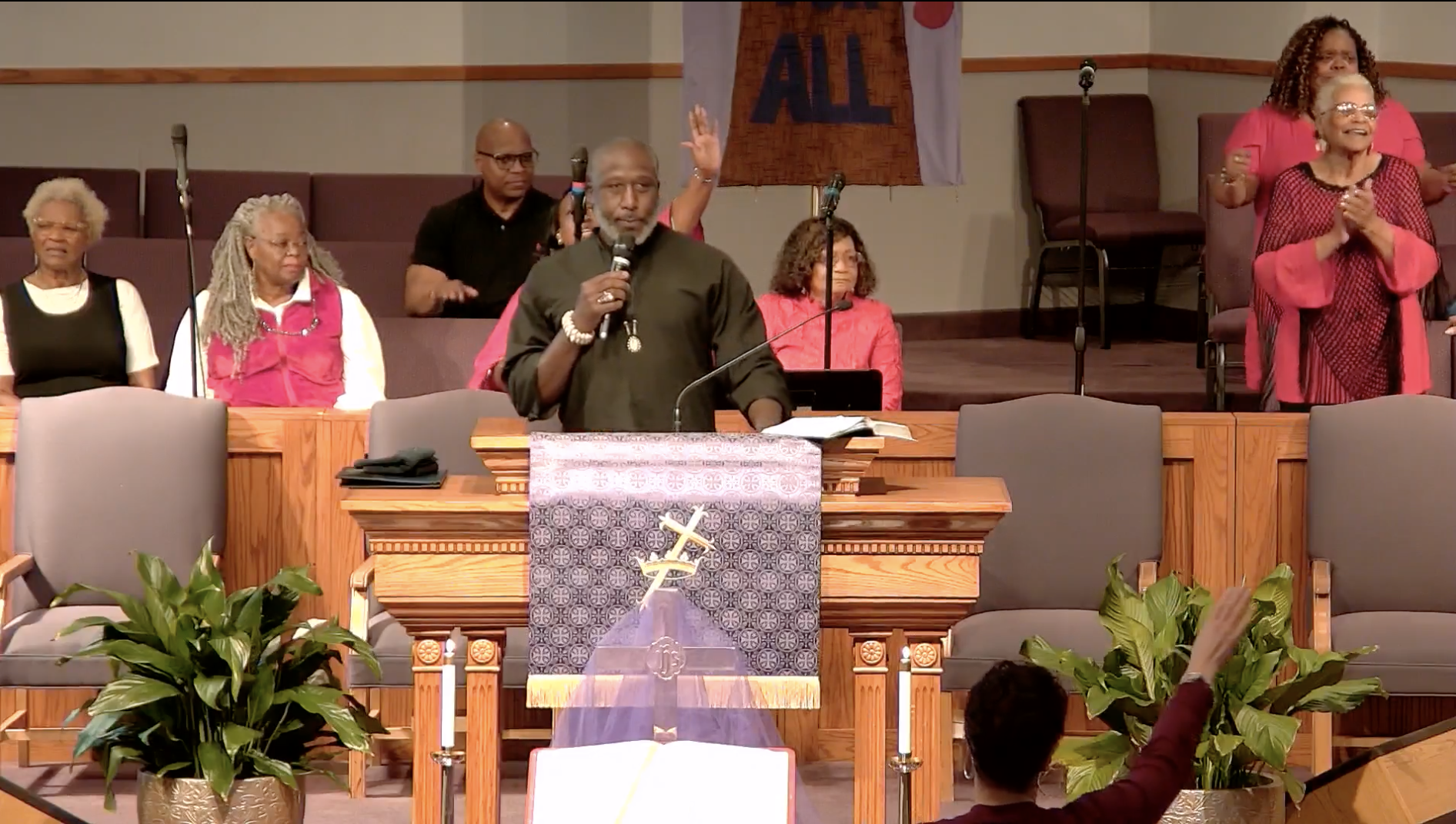 A diverse group of people standing and sitting in a church, with a man speaking at a pulpit and others clapping or raising their hands, indicating a religious gathering or service.