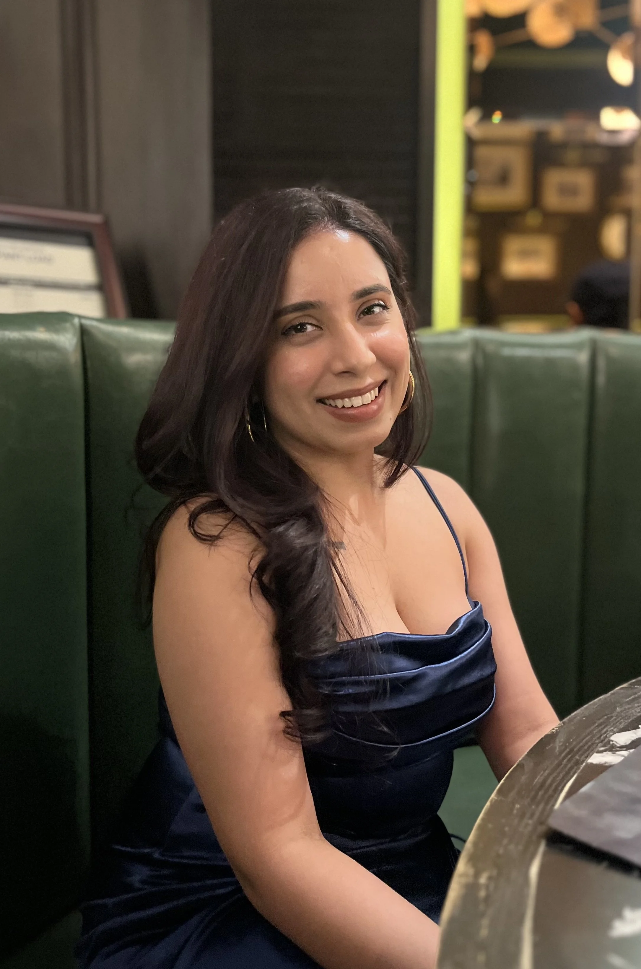 A smiling woman with long dark hair, wearing a navy blue satin dress, sitting on a green cushioned bench in a restaurant or cafe with a dark interior and warm lighting.
