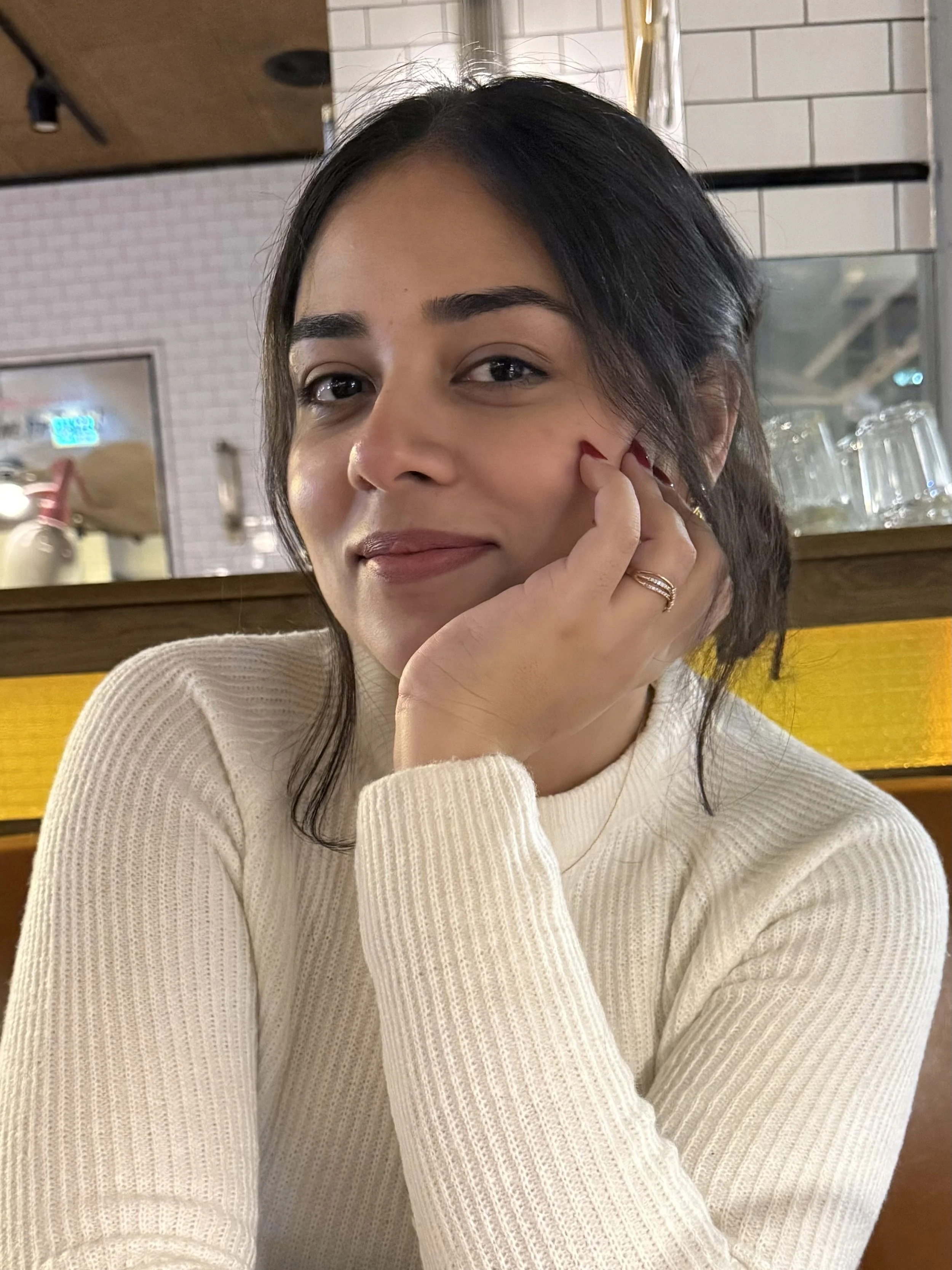 A woman with black hair and light skin is resting her chin on her hand, looking at the camera and smiling softly. She is wearing a cream-colored ribbed sweater, and her nails are painted red. The background includes white tiles and a wooden surface with glasses and metal fixtures.