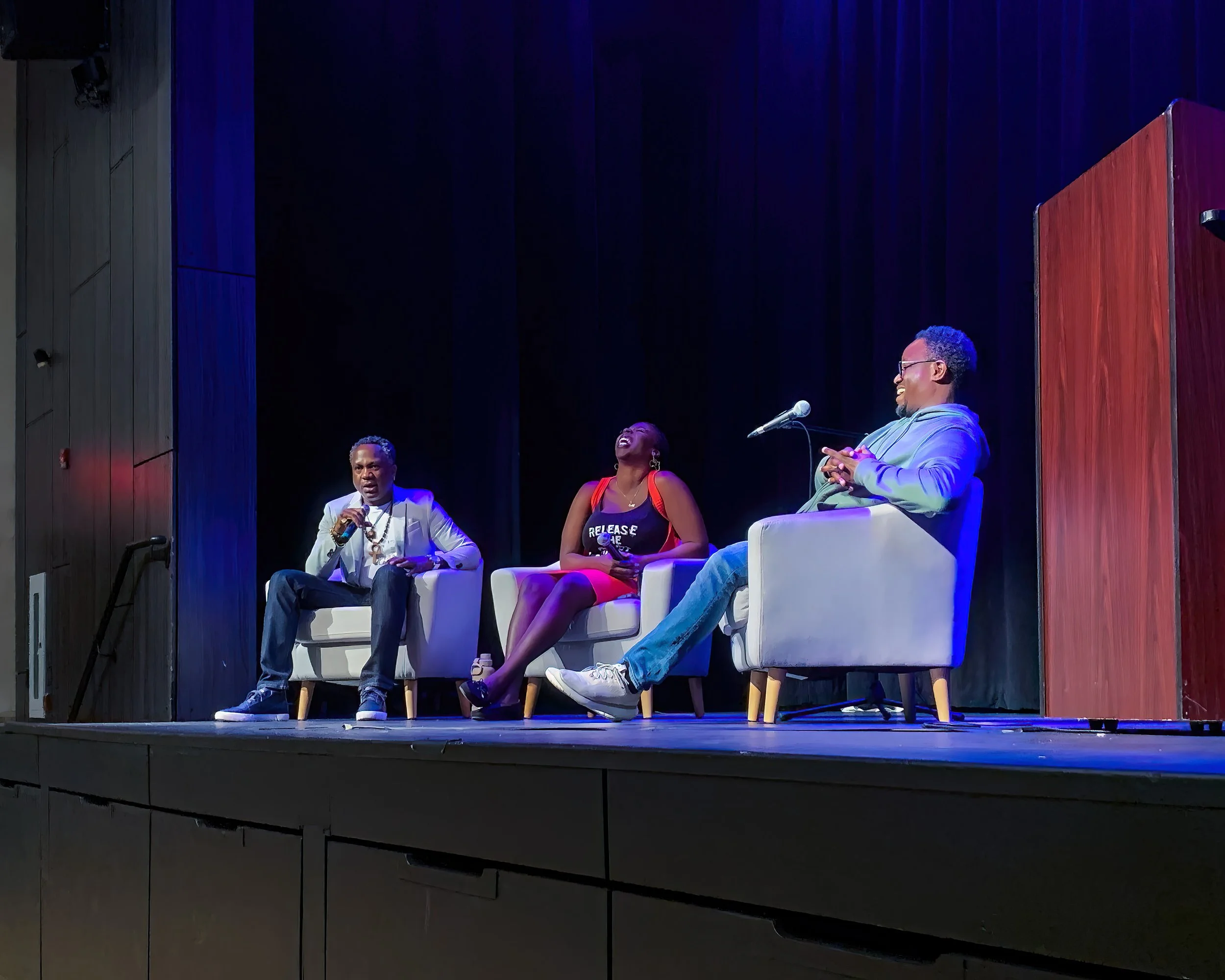 Three people sitting on stage in chairs engaging in a panel discussion, with a black curtain backdrop and stage lighting.