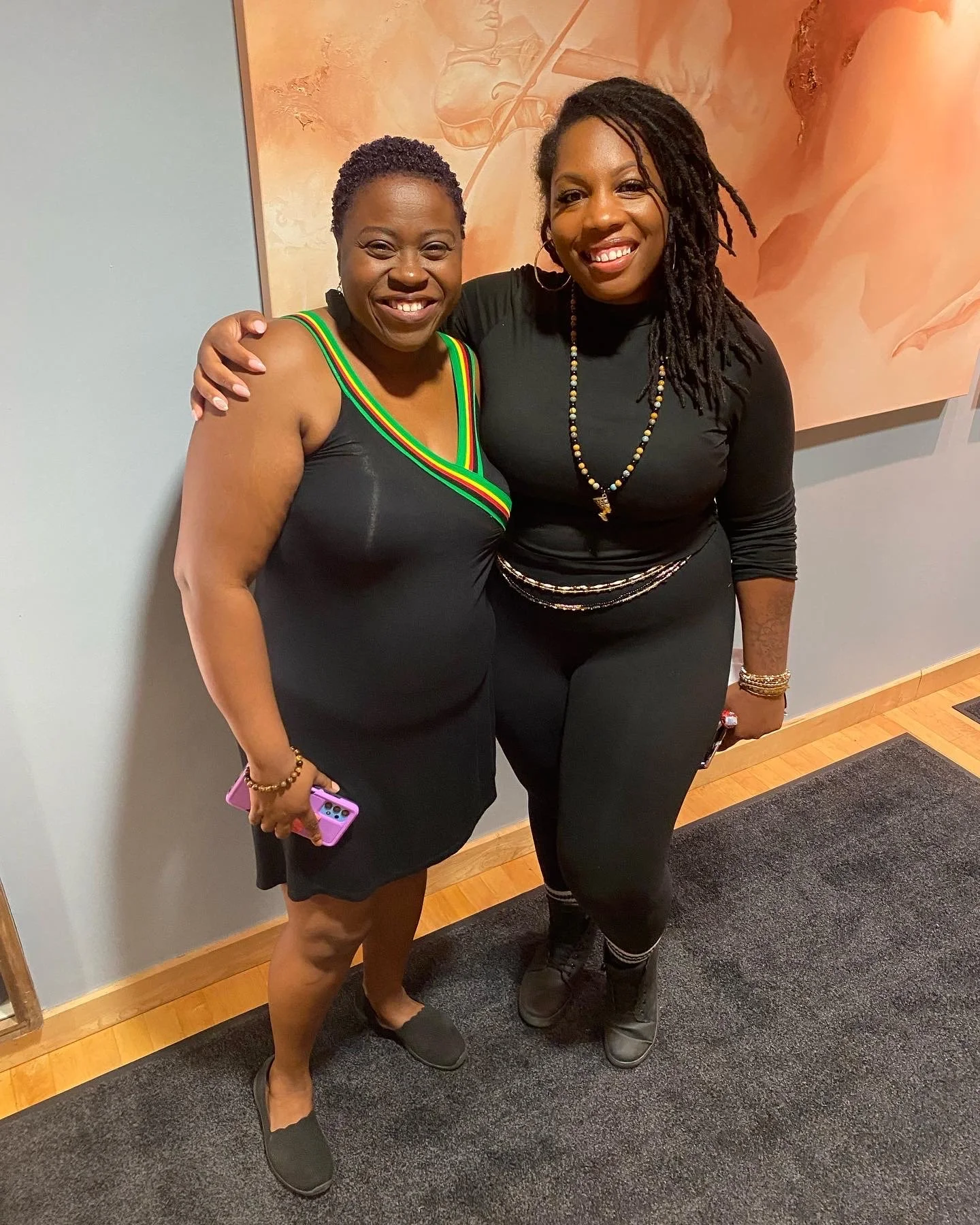 Two women standing side by side, smiling and posing for the photo. They are dressed in black clothing, one with a dress with green, yellow, and red stripe accents, and the other with a black top and pants. They are indoors, with a wooden floor and an