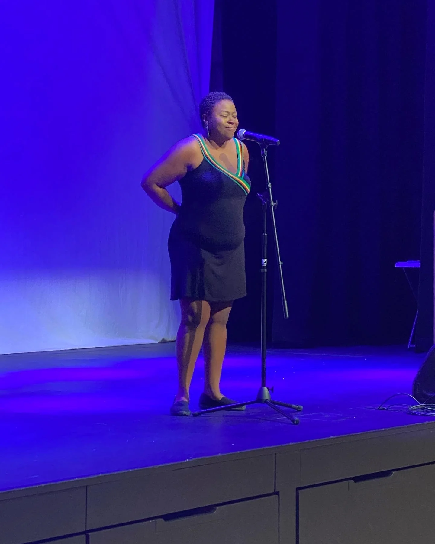 A woman in a black dress with rainbow-colored trim stands on a stage speaking into a microphone, with her hands clasped behind her back.