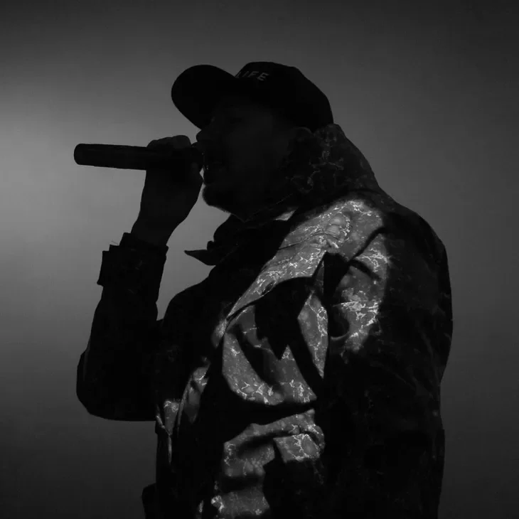 Silhouette of a person wearing a cap and jacket, holding a microphone, with a dark background.