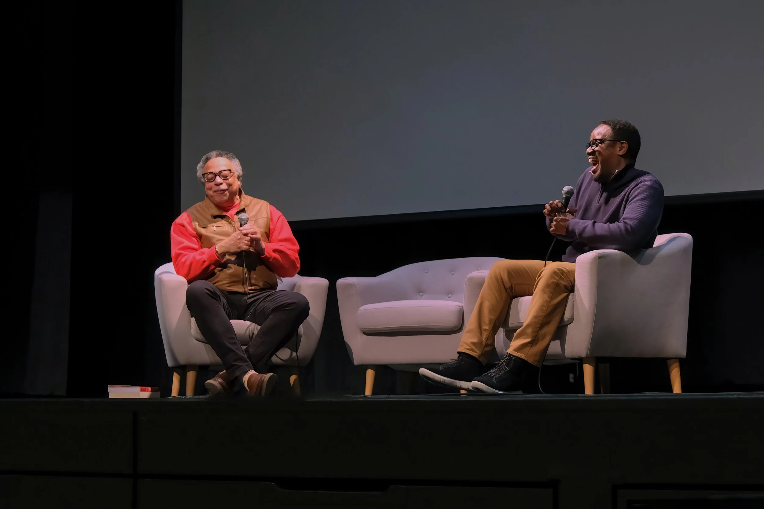 Two men sitting on stage in armchairs, engaging in a conversation with microphones, with a large blank screen behind them, smiling and laughing.