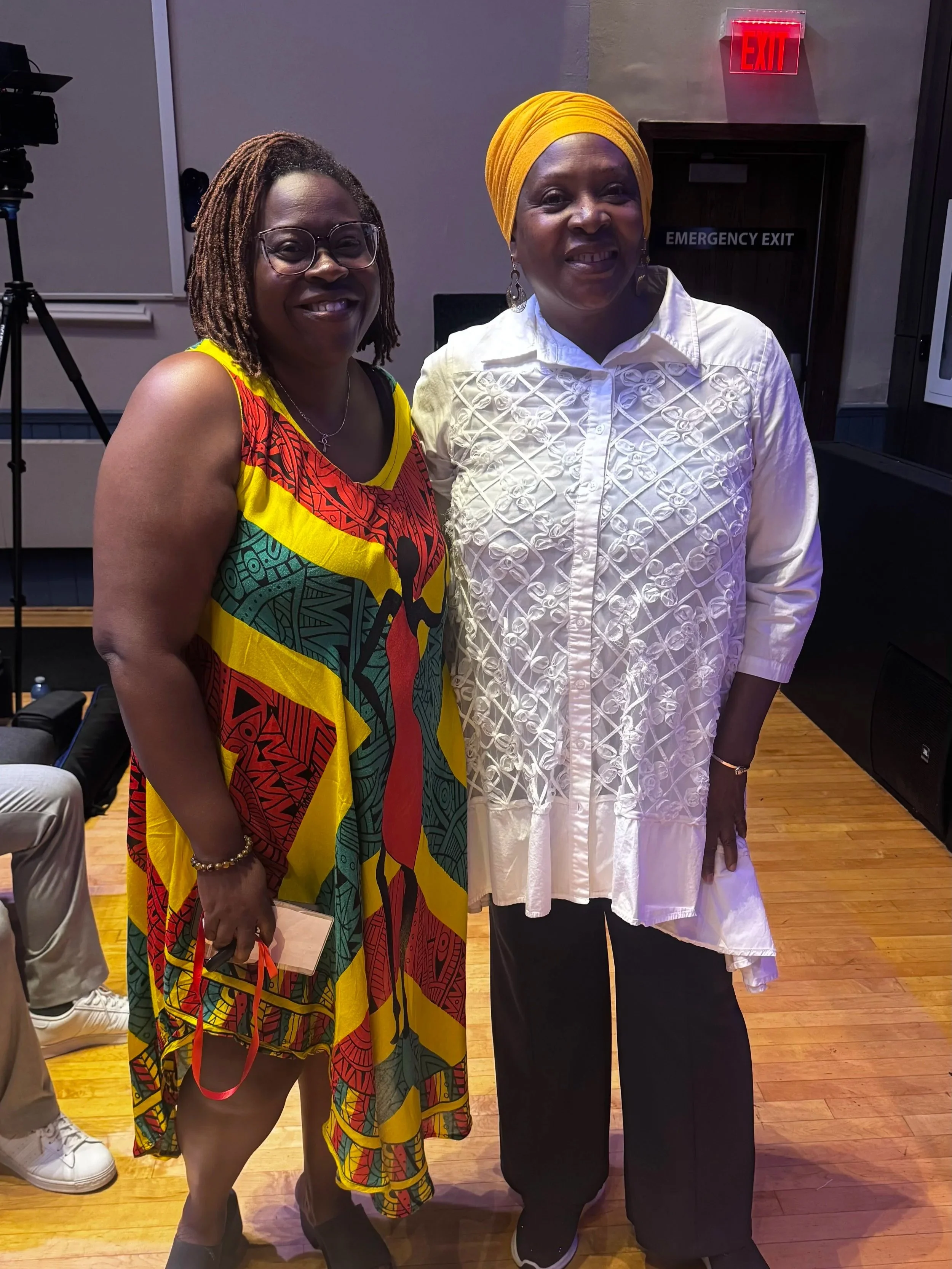 Two women standing together and smiling inside a room with a wooden floor. The woman on the left has shoulder-length dreadlocks, glasses, a colorful African dress, and is holding a pair of black shoes with red laces. The woman on the right is wearing a white embroidered dress, a yellow head wrap, large earrings, and has dark lipstick. There is a red exit sign and an emergency exit door behind them.