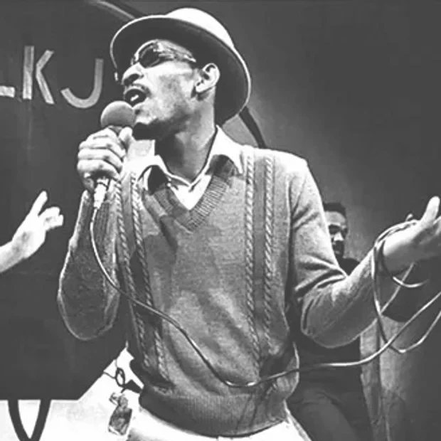 A man wearing sunglasses, a hat, and a sweater, holding a microphone and singing or speaking passionately on stage, with another person partially visible in the background.