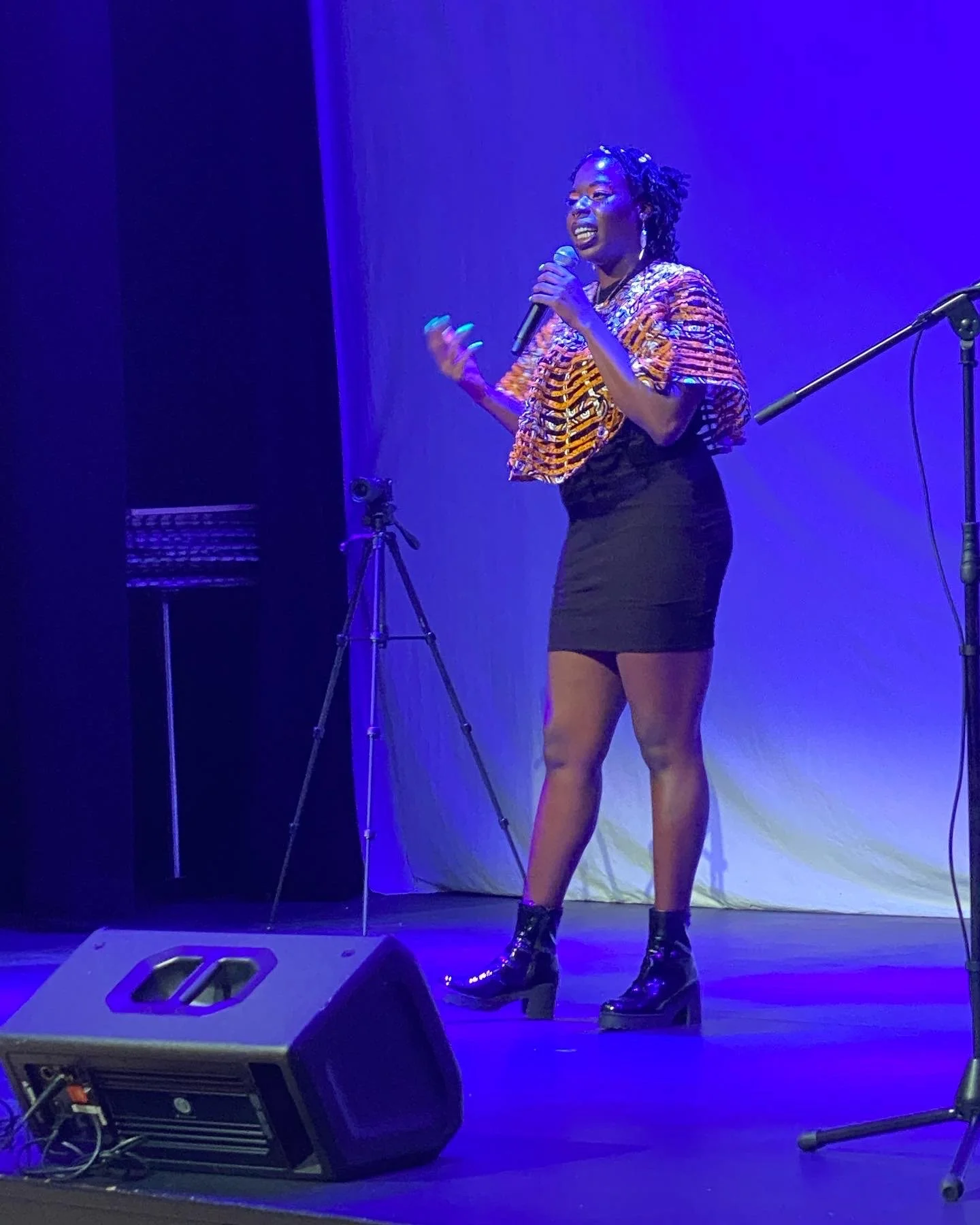 A woman standing on stage under blue lighting, holding a microphone, and singing or speaking. She is wearing a colorful striped top, a black skirt, and black ankle boots with heels.