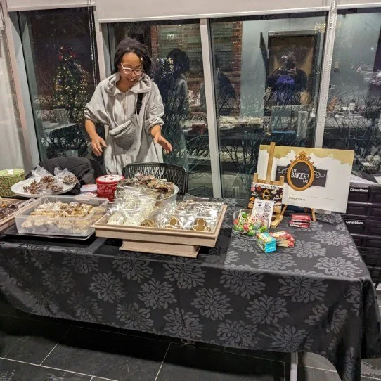A woman at a table filled with baked goods and candy, smiling while looking at the treats, next to a window decorated with a Christmas tree in the background.