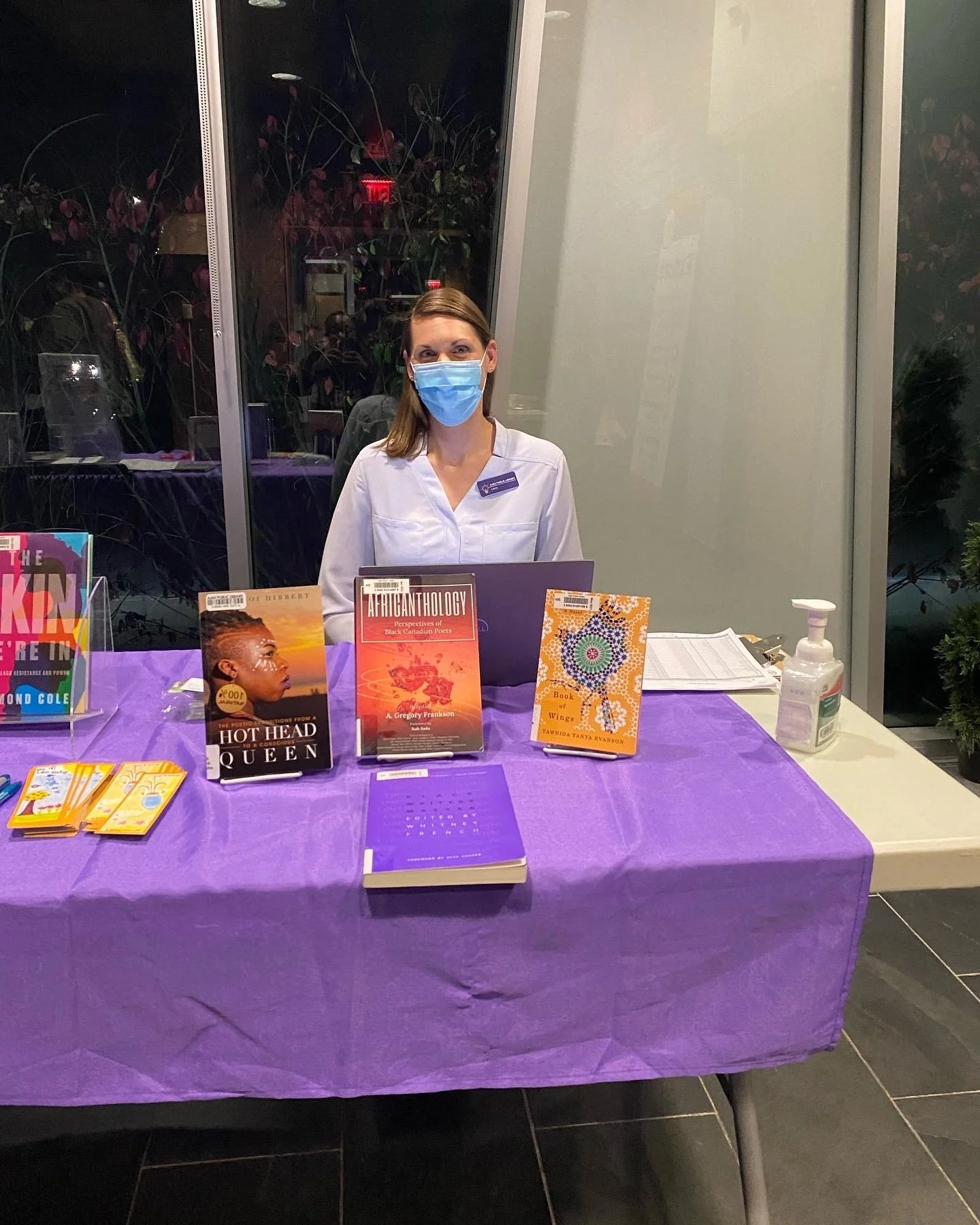 A woman wearing a blue face mask and white shirt sitting behind a table with purple tablecloth, books, and a hand sanitizer bottle, in a well-lit indoor setting.