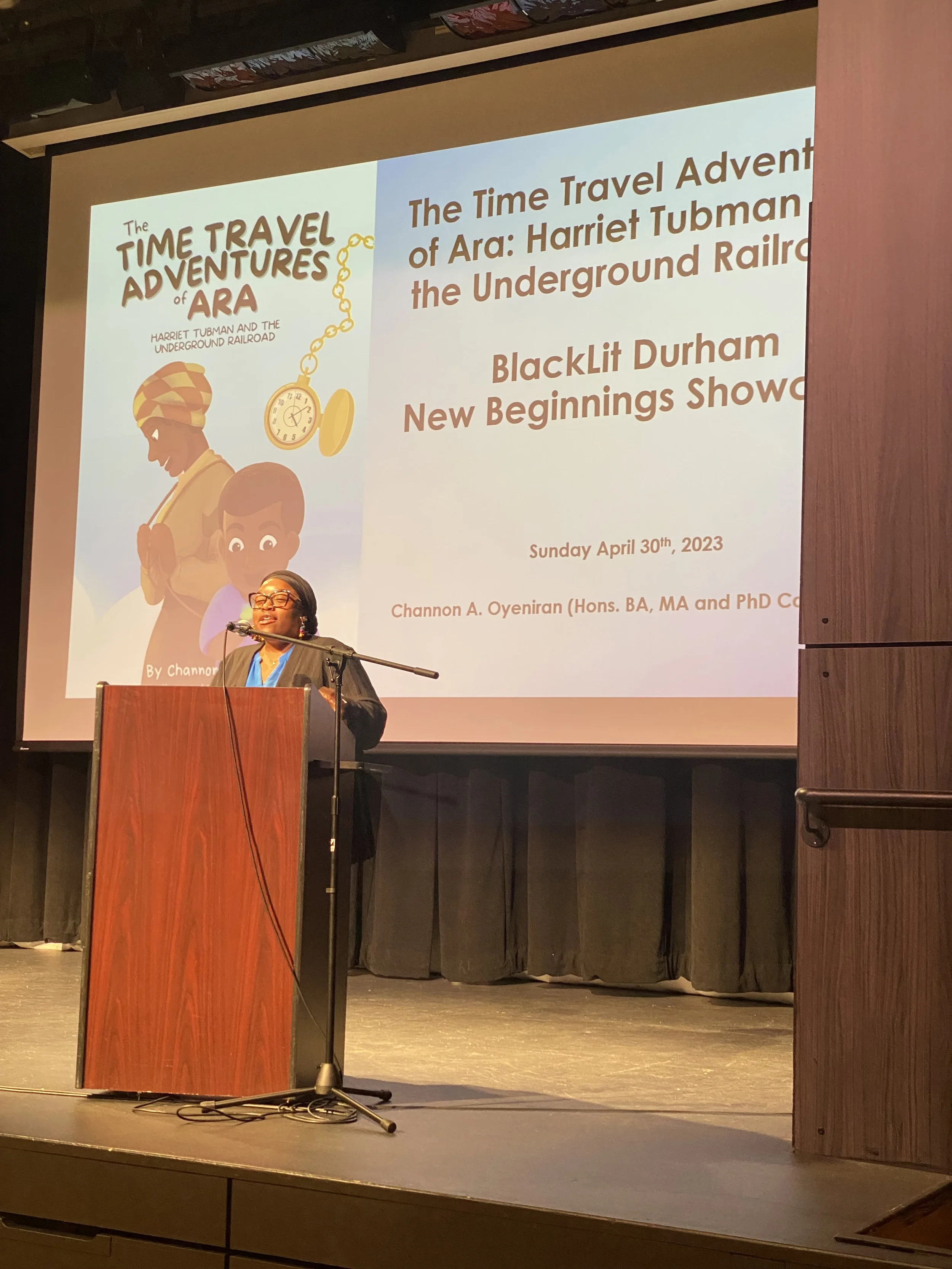 The children's writer Channon A. Oyeniran stands at a podium giving a presentation in a dark auditorium. Behind her, a large screen displays an illustrated book cover titled 'The Time Travel Adventures of Ara'.