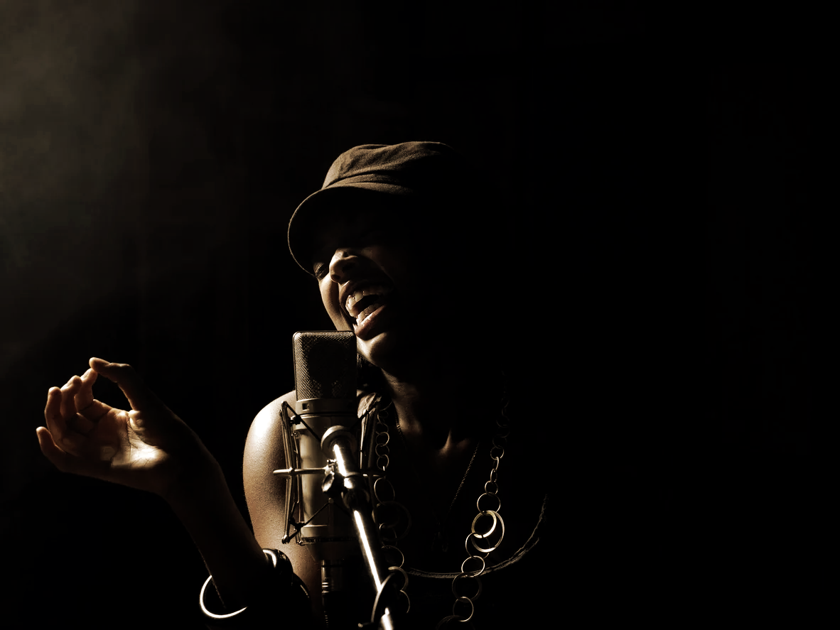 A person singing or speaking into a microphone in a dark studio, wearing a hat and large jewelry, with dramatic lighting highlighting their face and hand.