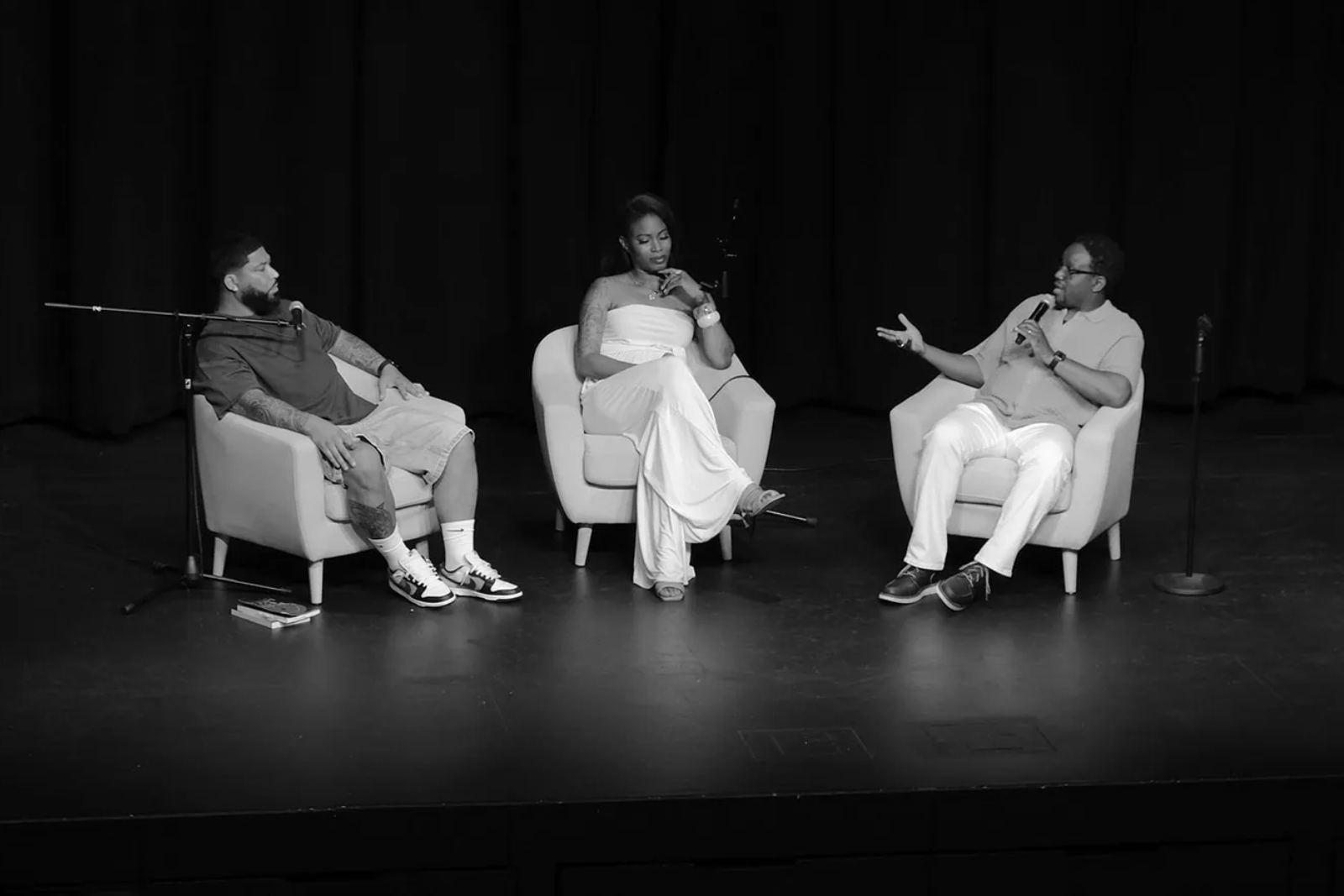 Three people sitting on stage in chairs having a discussion, with two men and one woman. All are holding microphones, and there are two additional microphones on stands nearby. The background is dark, and the scene is black and white.