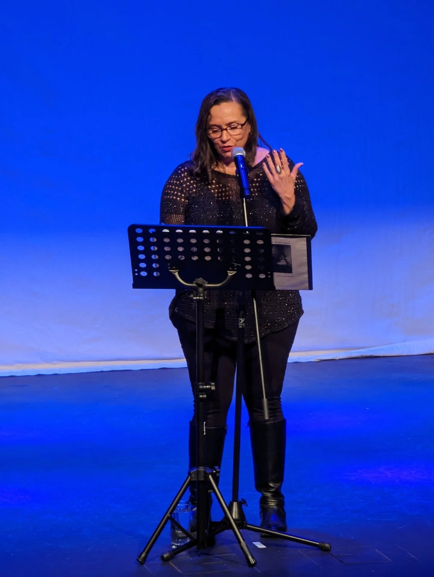 Woman with glasses and dark hair reading from a paper on a stand, speaking into a microphone on a stage with blue backdrop.