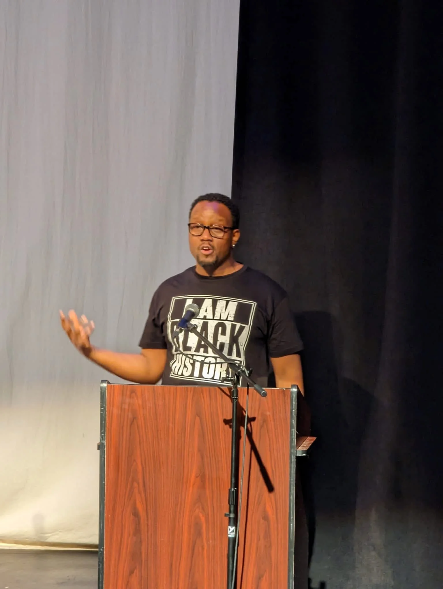 Man speaking at a podium on stage, wearing a black T-shirt that says 'I AM BLACK HISTORY,' with a microphone in front of him.