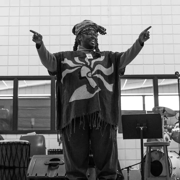 A person with long braided hair and glasses, wearing a bandana, black pants, and a flag with a lion emblem, standing with arms extended and pointing.