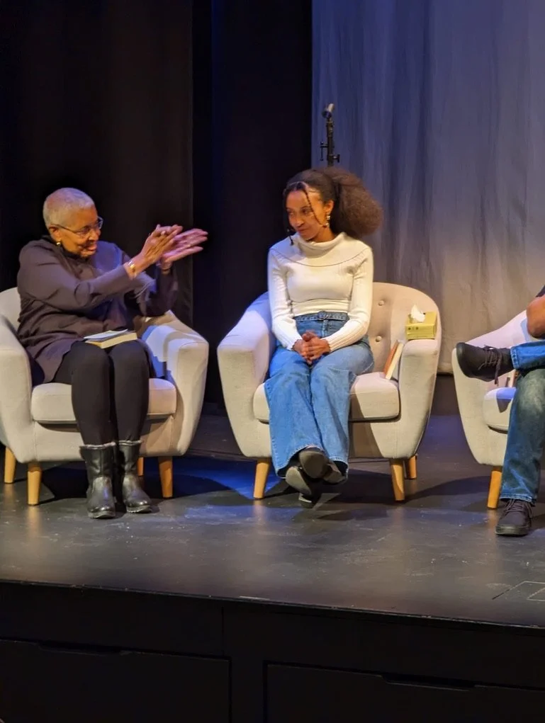 Three people sitting on beige armchairs on a stage, engaged in conversation. The person on the left is an elderly woman clapping and smiling, wearing glasses and black boots. The person in the middle is a young woman with curly hair, wearing a white