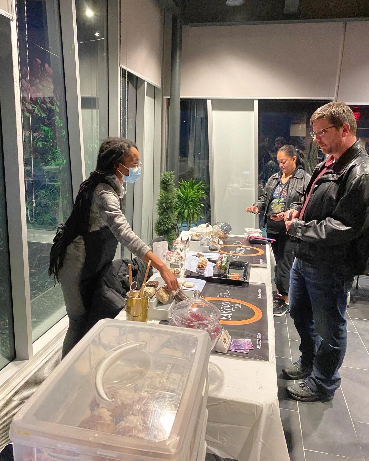 A woman wearing a mask and black apron serves baked goods at a table, while two customers, a man and a woman, stand in front of the table. The table is set up with various baked items and informational materials in a public hall setting.