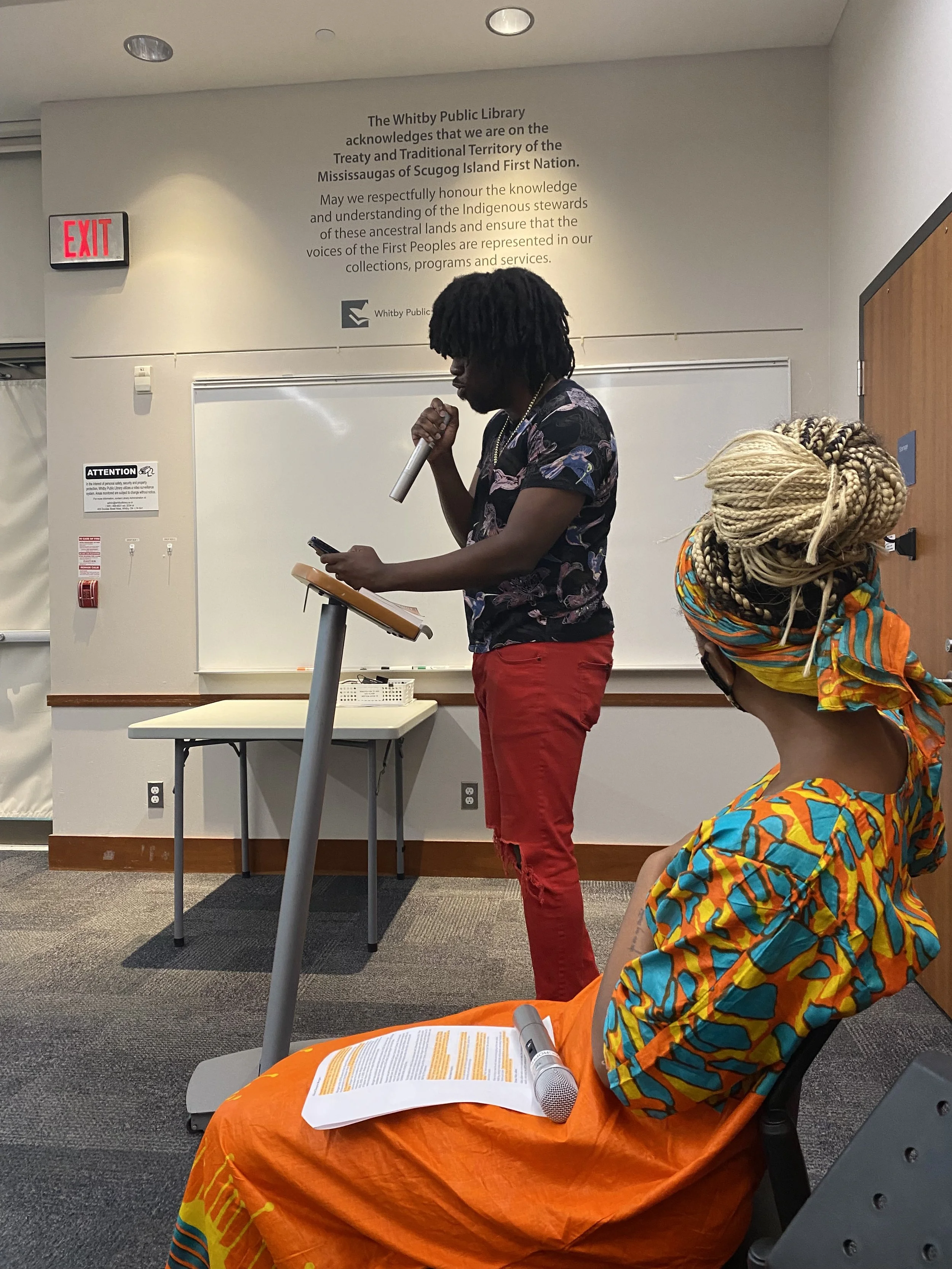 A speaker standing at a podium, reading from a phone, while a woman dressed in vibrant traditional African clothing and head wrap sits in front, holding a sheet of paper and a microphone. The setting is an indoor event at the Whitby Public Library.