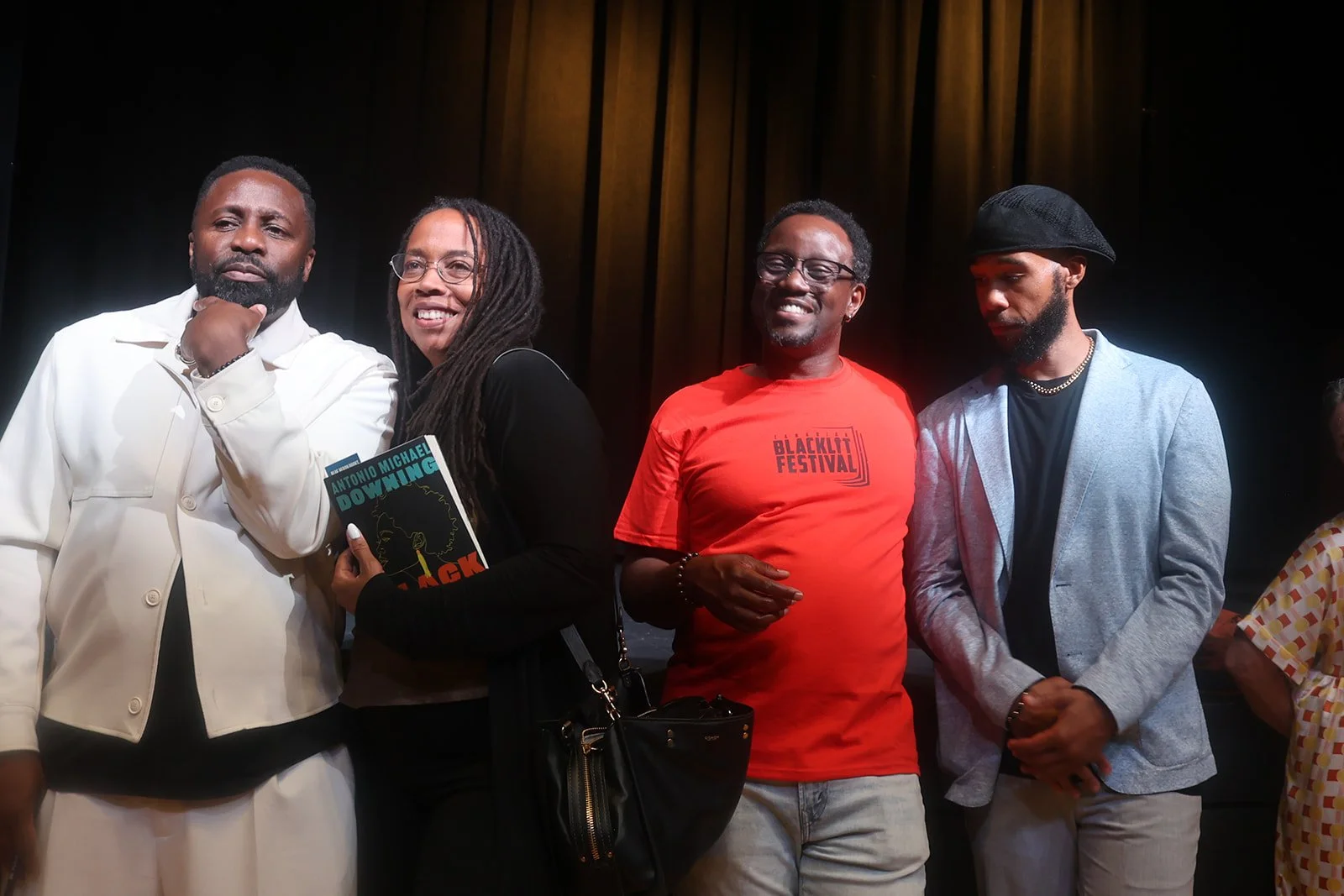 Group of four diverse people standing on a stage, smiling, with a dark curtain background, one holding a book titled "Antonio Michael Downing - Black" and wearing a black jacket and glasses, another wearing a red "Blacklit Festival" t-shirt, and others in casual and contemporary attire.