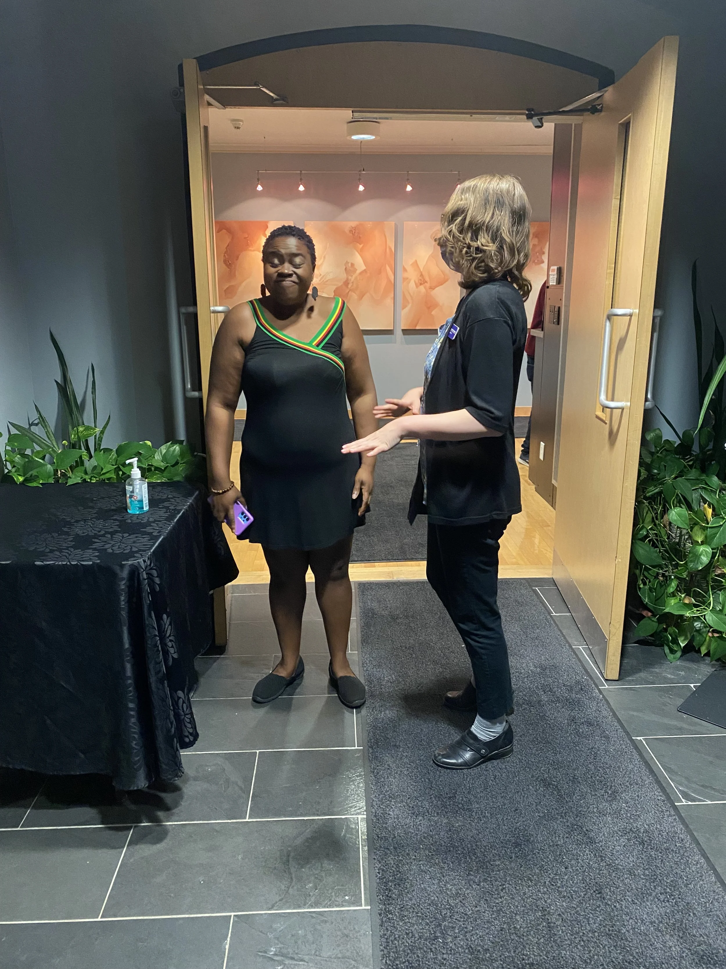A woman in a black dress with colorful stripes around the neckline is greeted by a staff member at the entrance of the St. Francis Centre, with hand sanitizer on a table nearby.