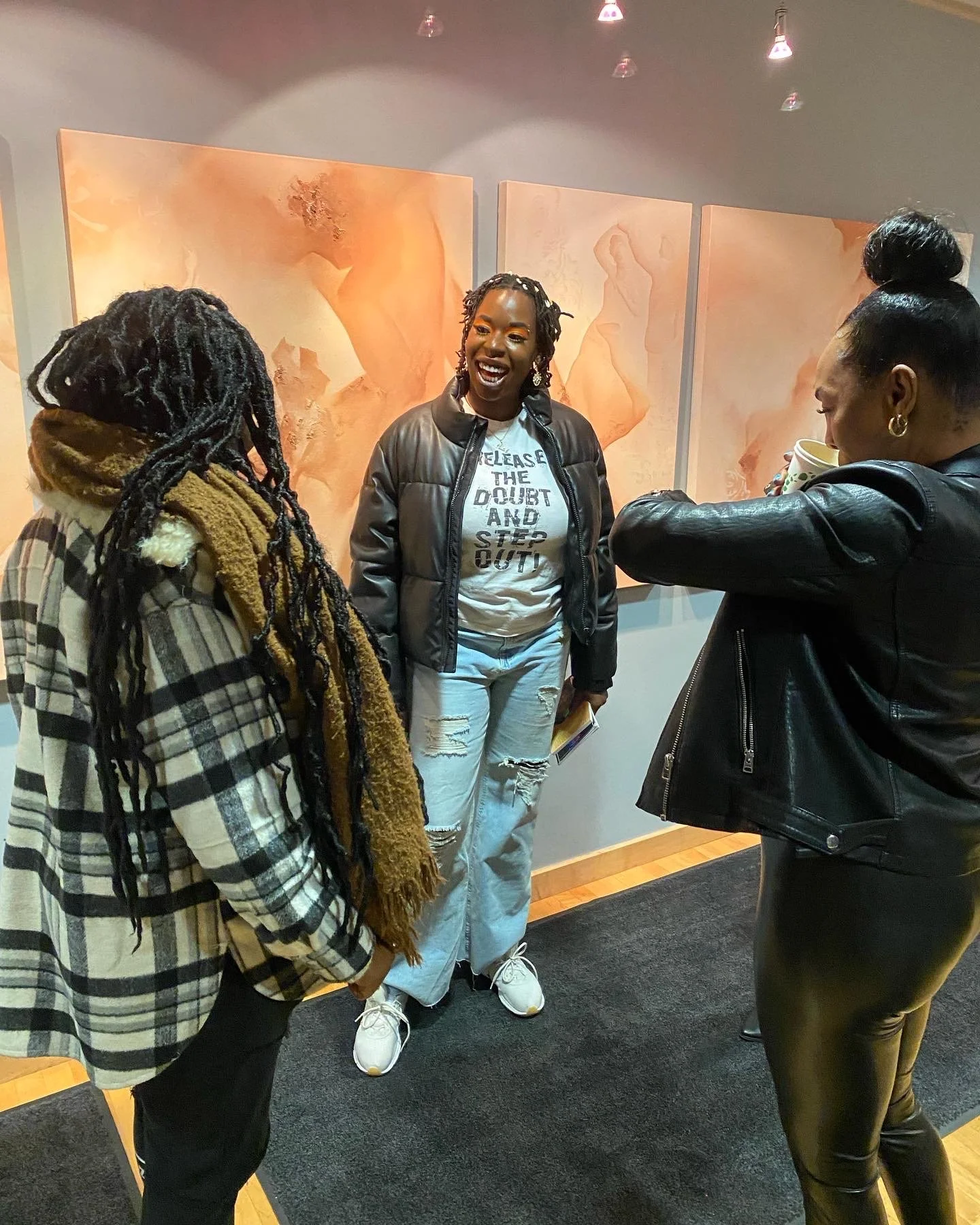 Four women talking and laughing in an indoor art gallery with three large paintings of abstract female forms in soft peach tones on the wall behind them.
