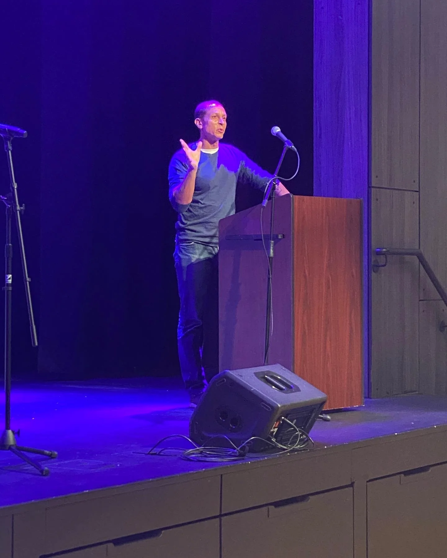 A person standing at a wooden podium on a stage with purple ambient lighting, speaking into a microphone, with a speaker on the floor and some cables visible.