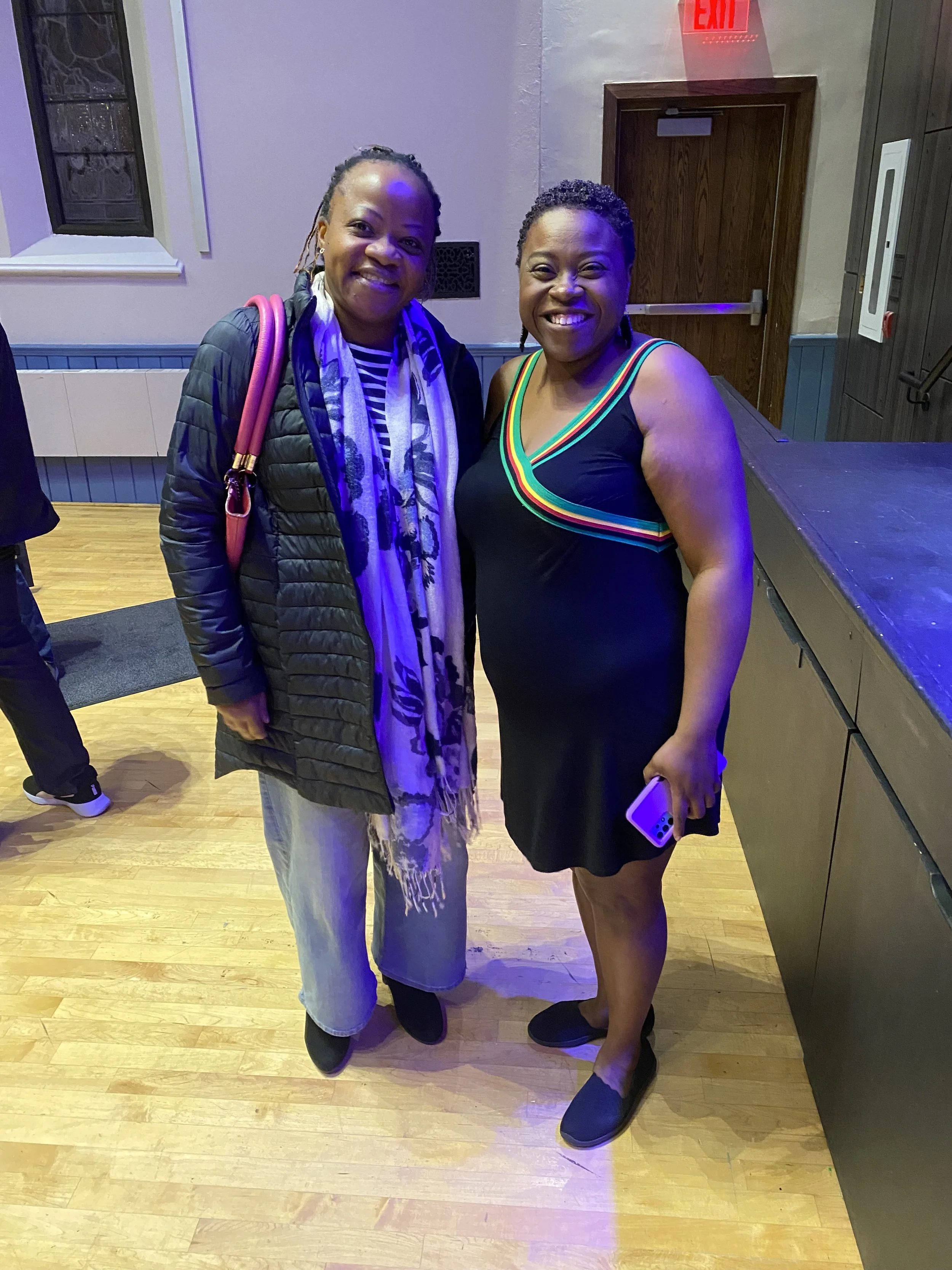 Two women standing side by side and smiling indoors, with wooden flooring and a wall with a window and a door behind them, one wearing a black dress with rainbow-colored straps and holding a phone, the other wearing a black quilted jacket, a striped