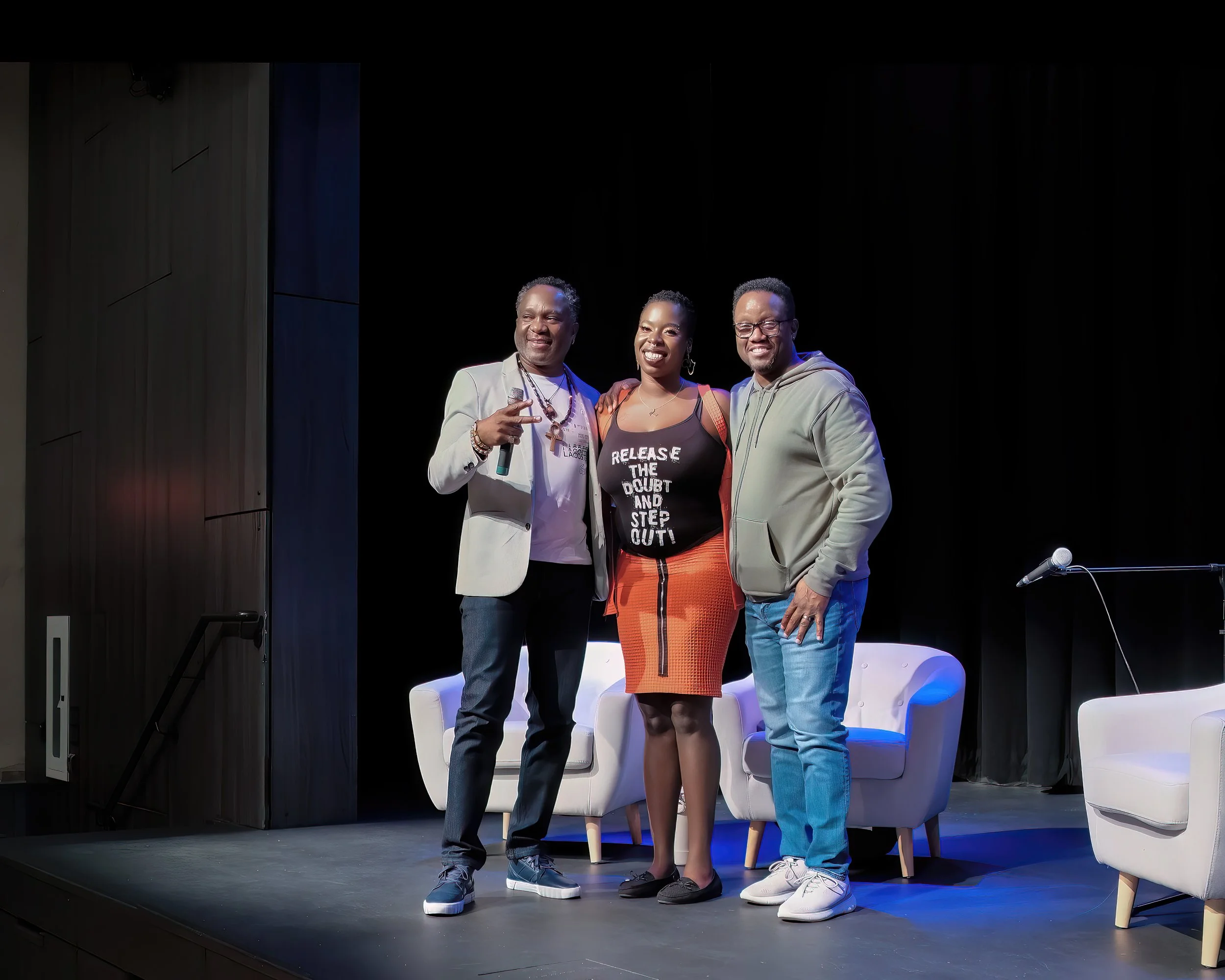 Three people standing on a stage with chairs, smiling for the camera. The person on the left is holding a microphone and making a peace sign. The person in the middle is wearing a black shirt with white text and an orange skirt. The person on the rig