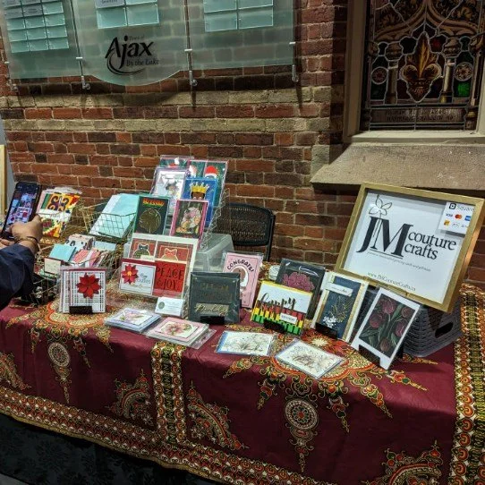 Display of handmade cards and crafts on a table covered with a patterned red cloth, set against a brick wall with a window.
